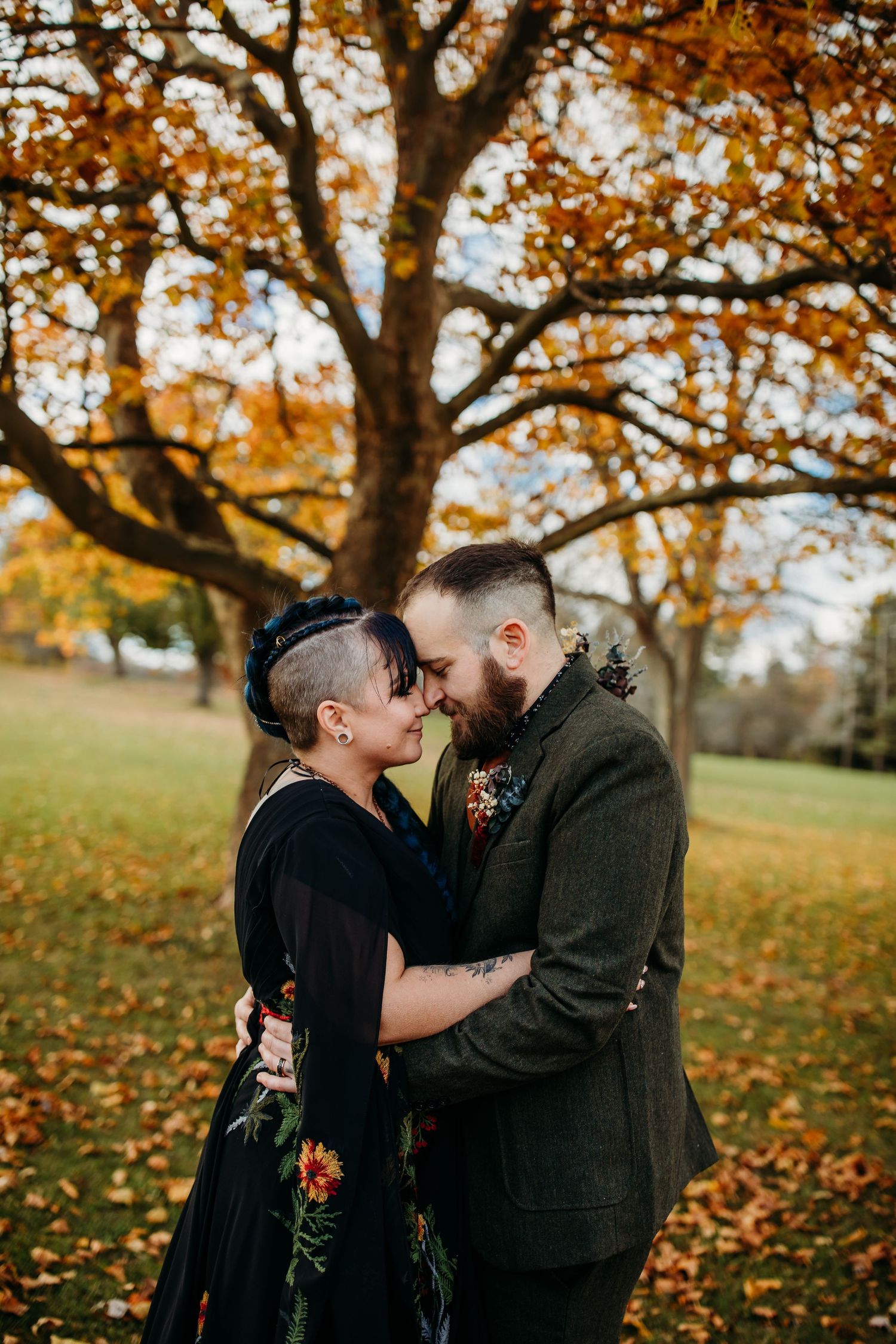 A queer couple embrace on their wedding day. The bride is wearing a black floral dress, and the groom has a brown suit on.