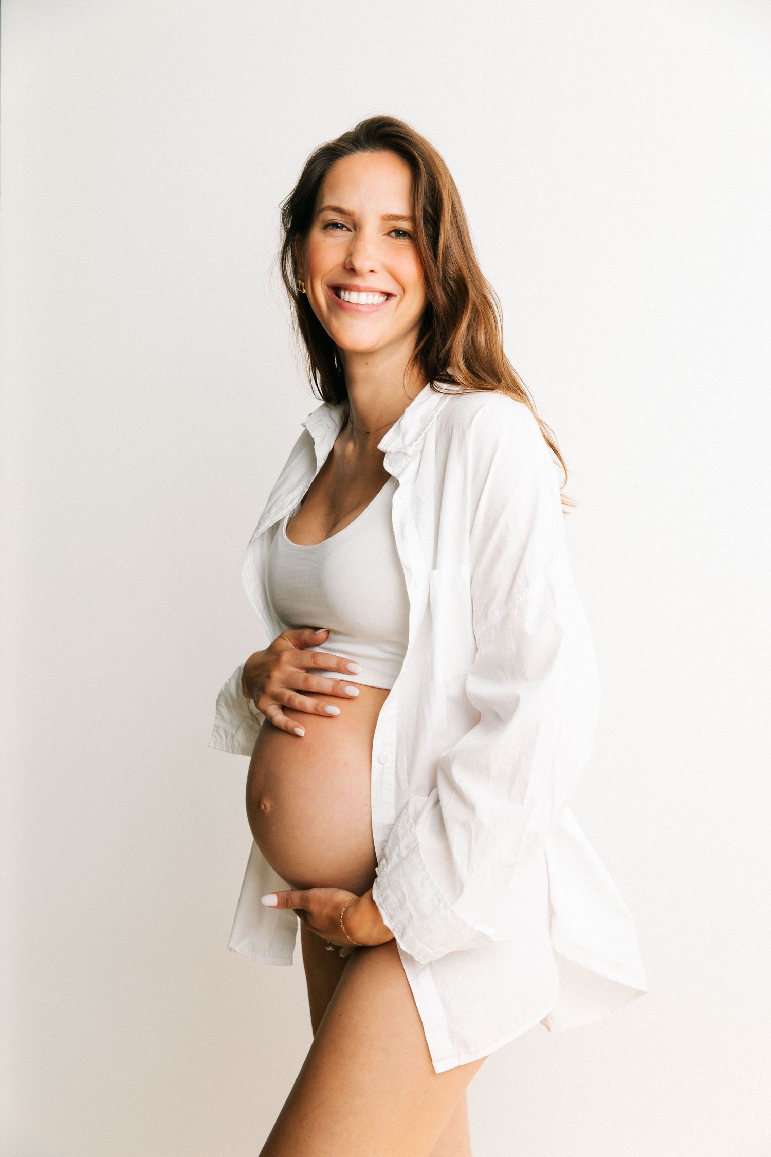 Creative maternity portrait of pregnant woman in oversized white shirt hands on belly smiling in Santa Monica studio by Phia Photography.