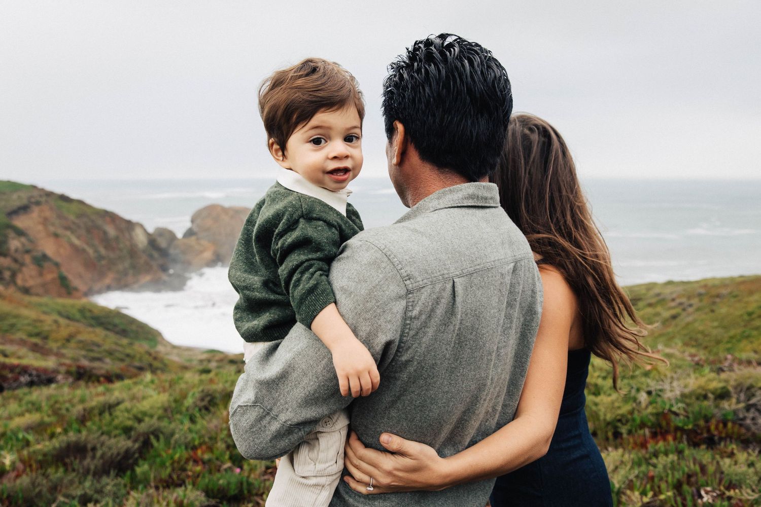 Candid family photo with parents looking over the natural beauty of Marin County coast and the child looking back at the photographer.