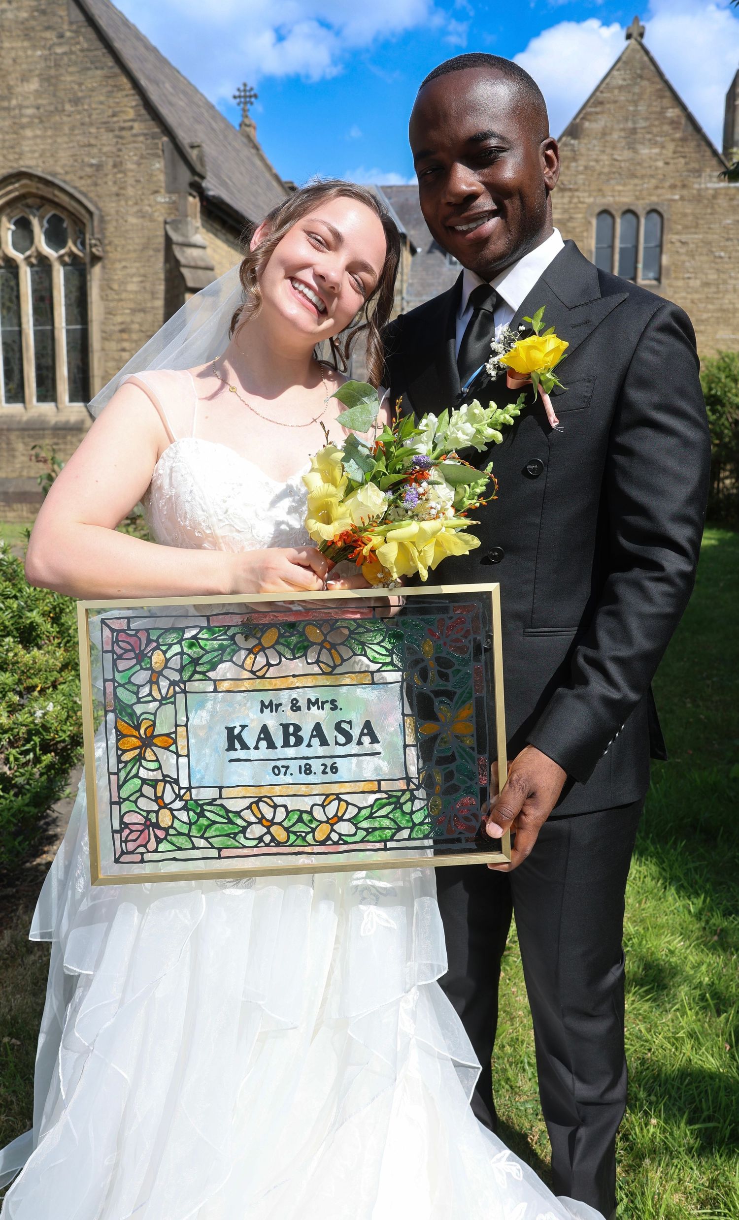 Mr. & Mrs. Kabasa pose in wedding attire outside a church, holding a stained glass sign dated 07.18.26.