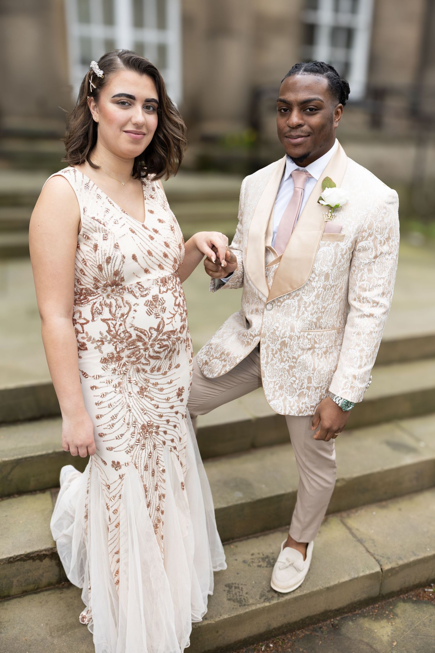 A couple poses on stone steps wearing coordinated cream and blush formal attire with delicate floral patterns.