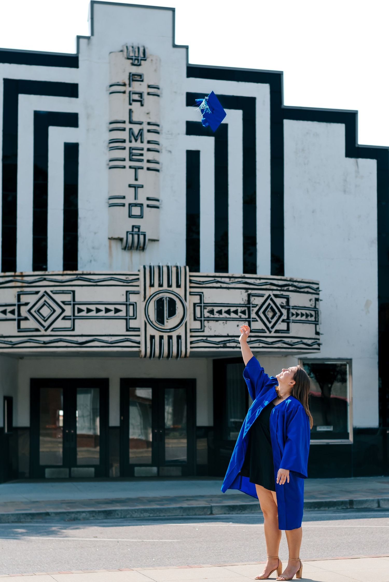 High school senior girl in bright blue grad gown tossing cap into air in front of theater in downtown Hampton, South Carolina.