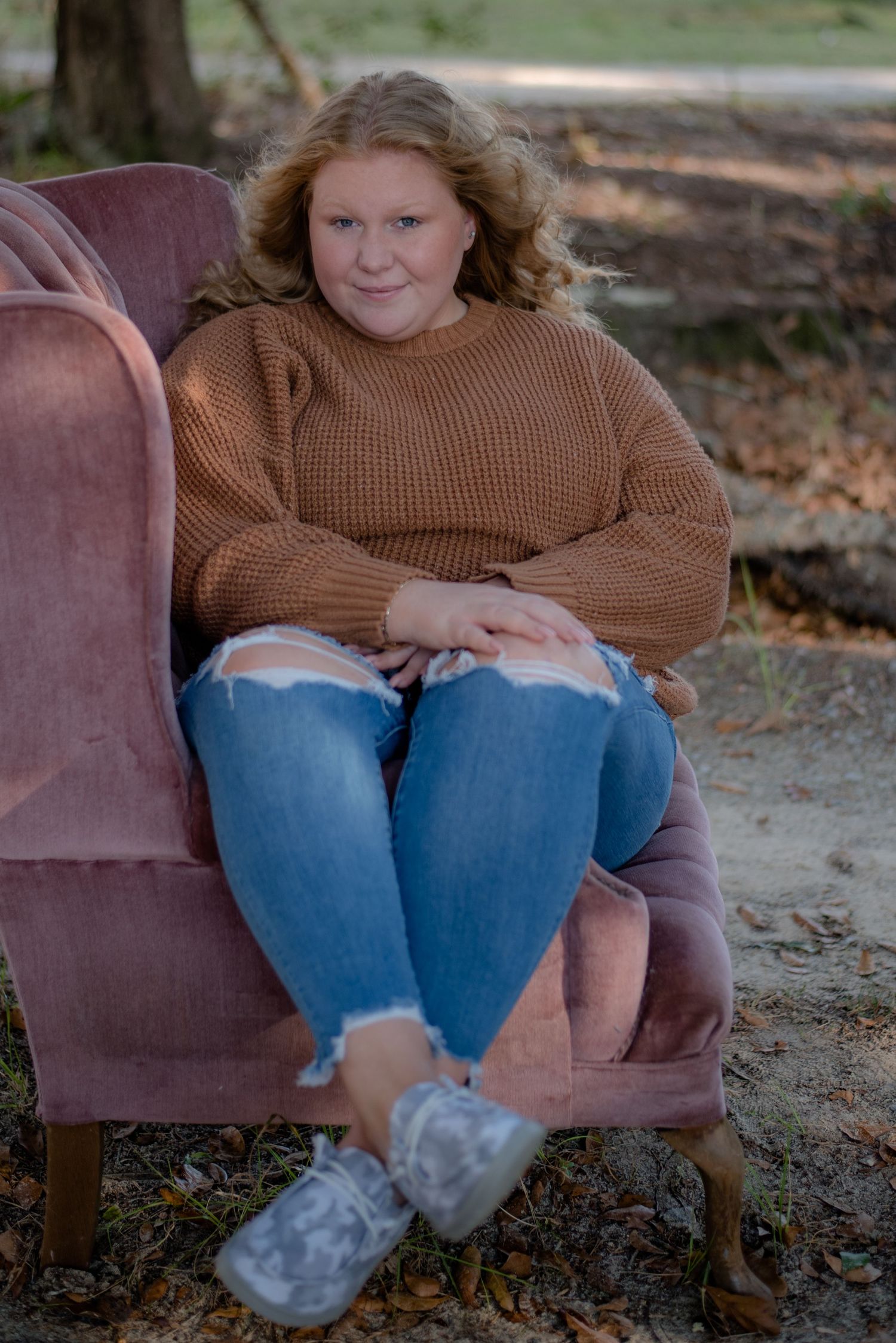 High school senior girl in fall, wearing ripped jeans and a sweater, sitting in a pink chair on gravel driveway in Hampton County, SC