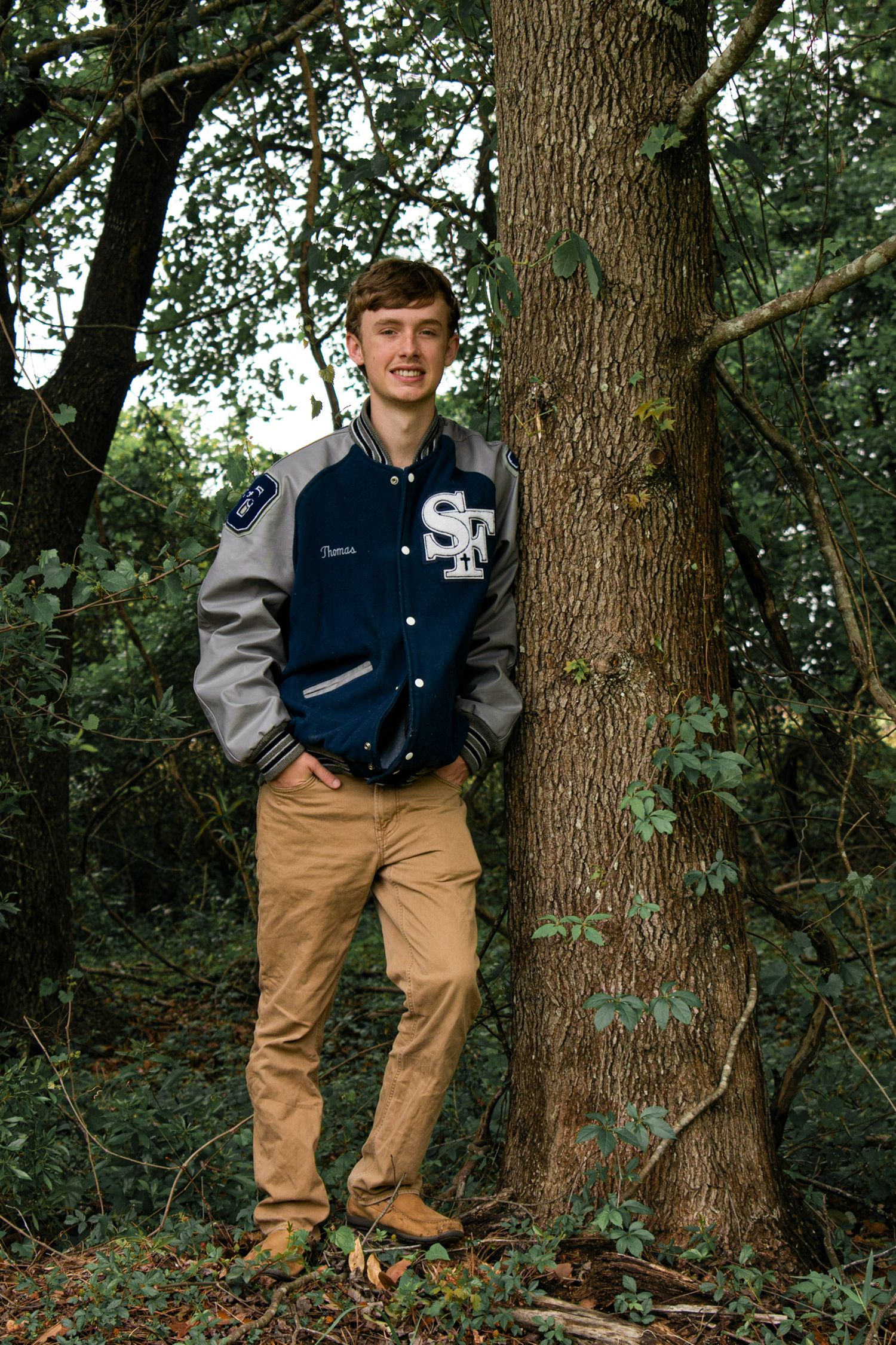 High school senior guy leaning against tree in letterman jacket in yard in Beaufort, South Carolina.
