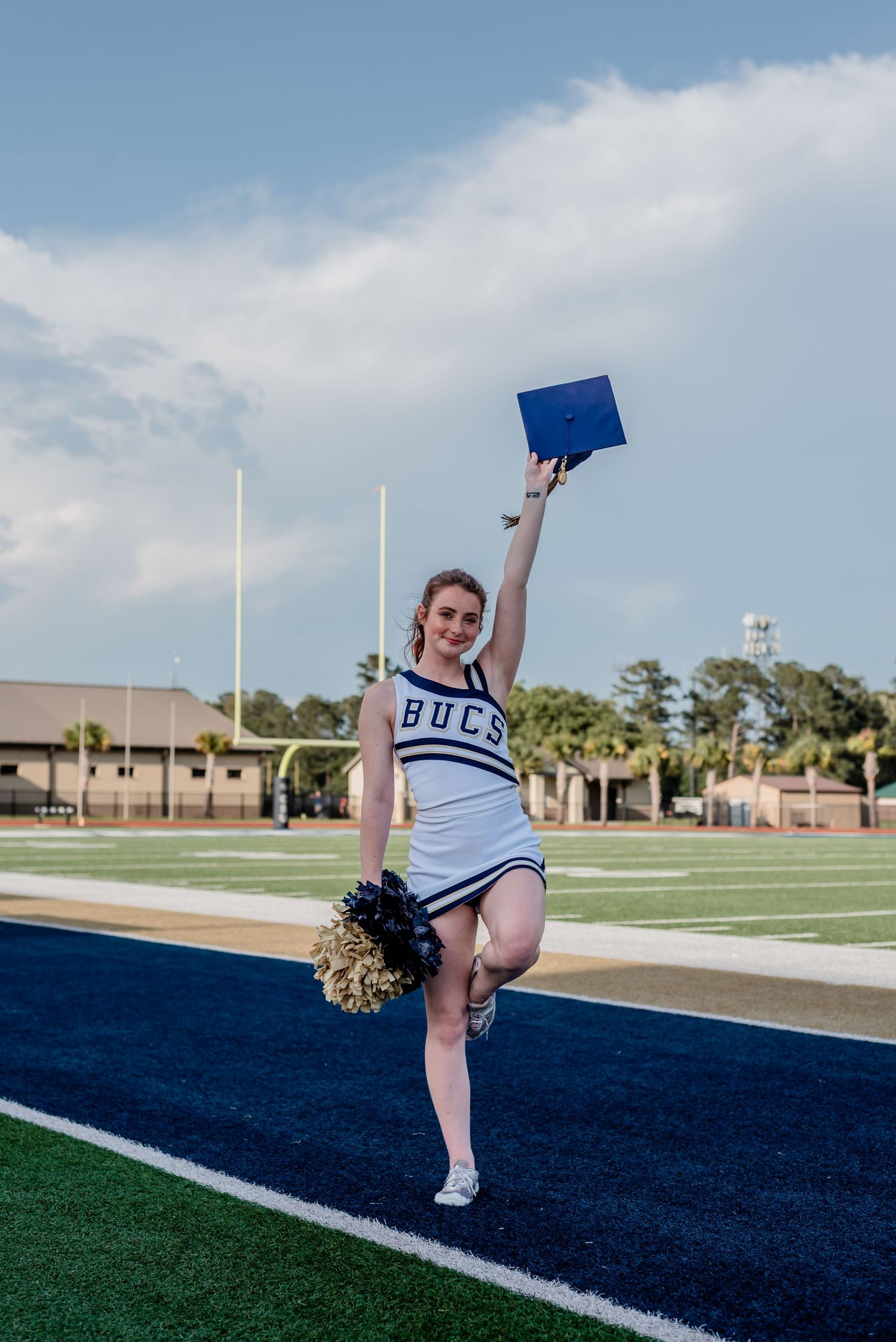 Charleston Southern University graduate wearing cheer uniform, holding pom-poms and graduation cap on football field in Charleston, SC.