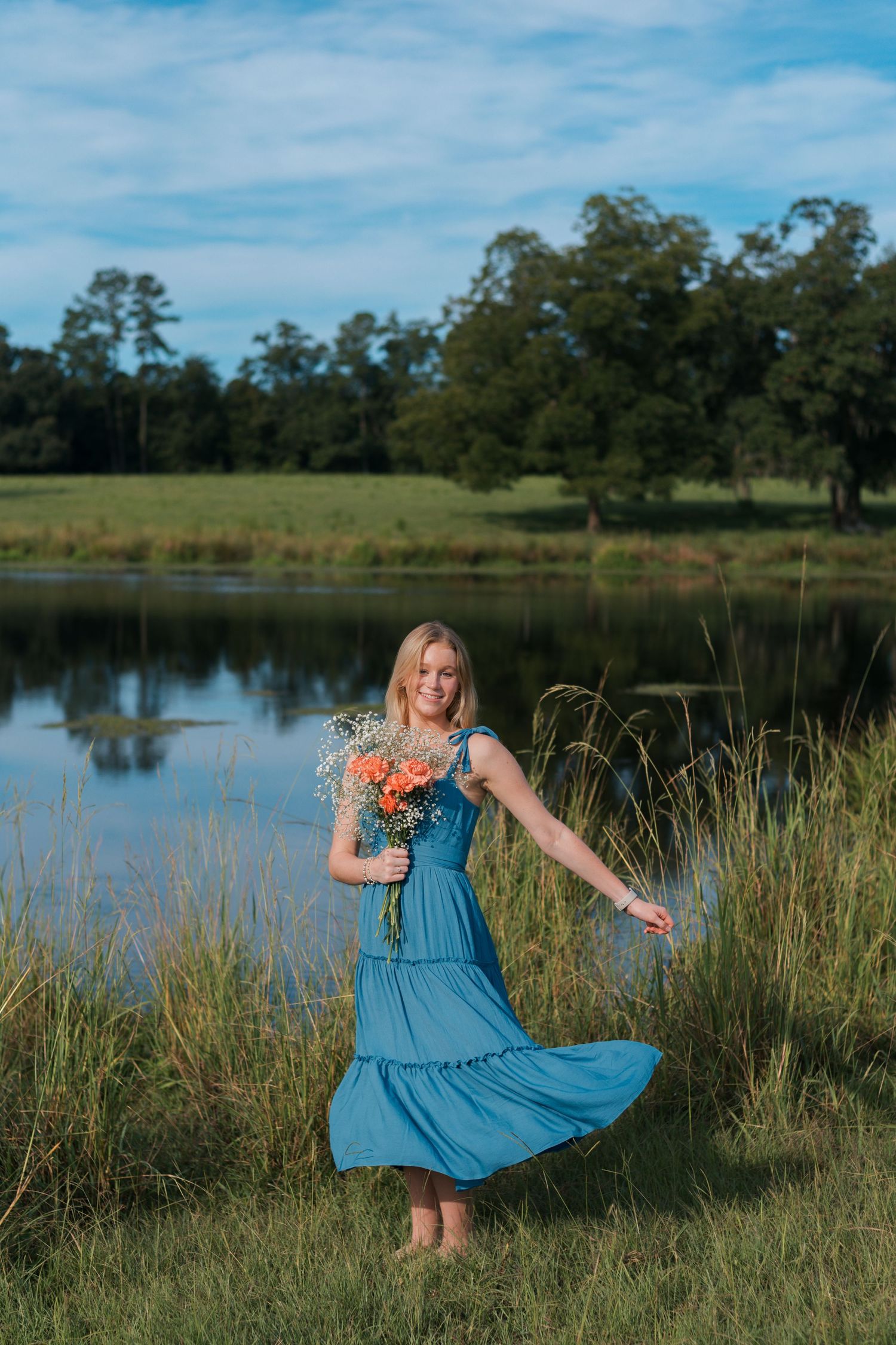 High school senior girl holding wildflower bouquet in blue dress in front of cow pond with large tree in the distance Farm in South Carolina