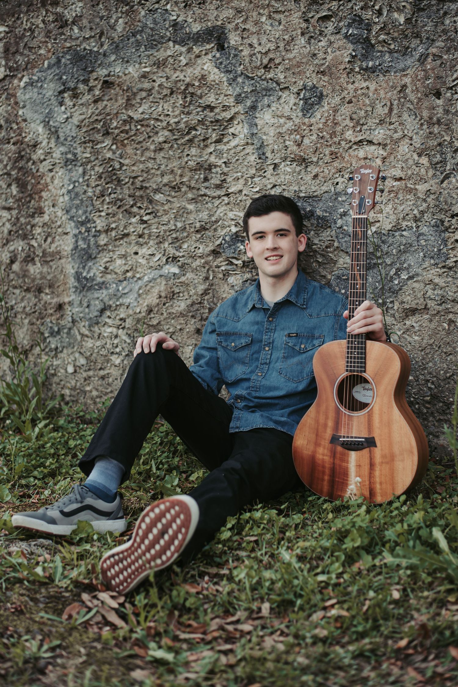 High school senior guy holding guitar, sitting in front of wall at Colonial Dorchester State Historic Site in Summerville, South Carolina.