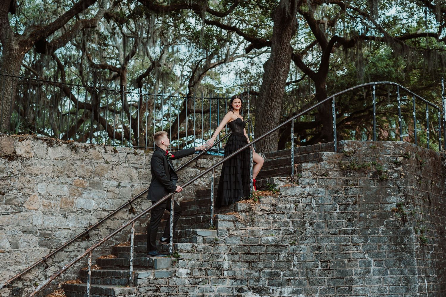 Couple wearing prom attire, black dress, tux and red accessories, climbing old steps in waterfront district of Savannah, Georgia
