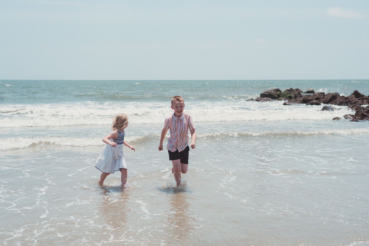 brother and sister running through ocean water with rocks in the background on Edisto Beach Edisto Island, South Carolina