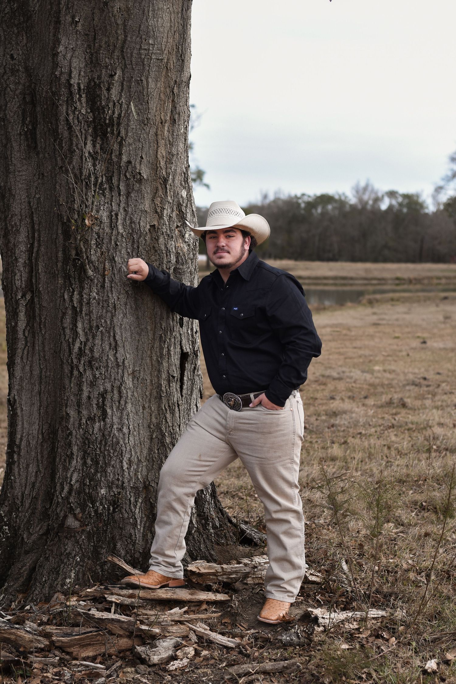 High school senior boy in cowboy boots, propped against tree in the middle of a pasture on a farm in Auburn, Alabama.