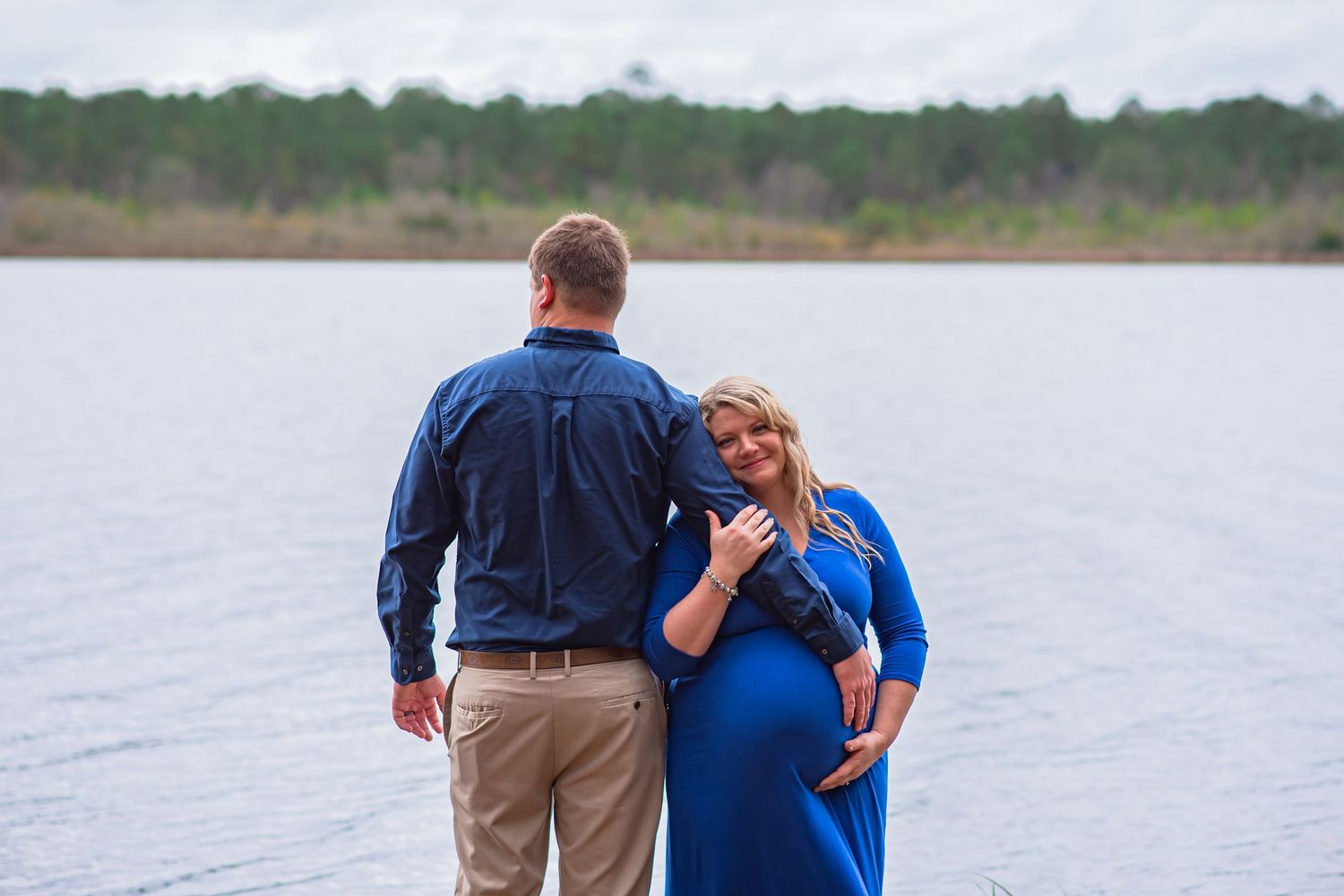 Husband facing Lake Warren while embracing pregnant wife's belly who is face the camera. Wearing shades of blue under winter sky. Hampton SC