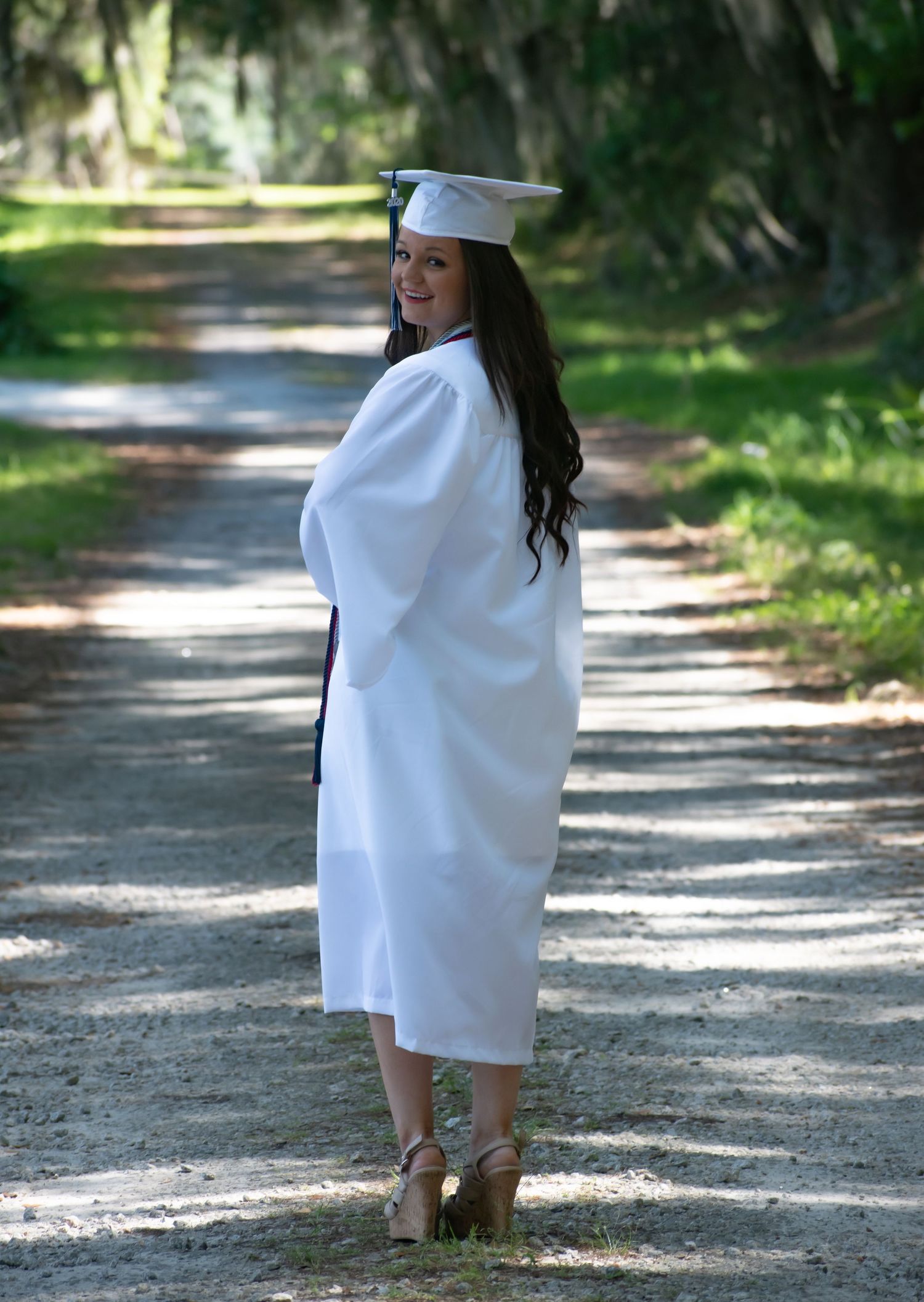 High school senior girl in white cap and gown wearing wedges on a gravel driveway at Thomas Heyward Historical Site in Ridgeland, SC