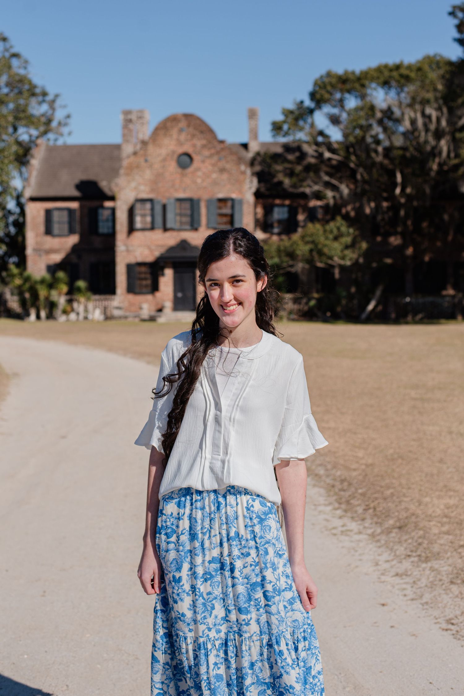 High school senior girl in long blue skirt in front of house on dirt driveway at Middleton Place in Summerville, South Carolina.