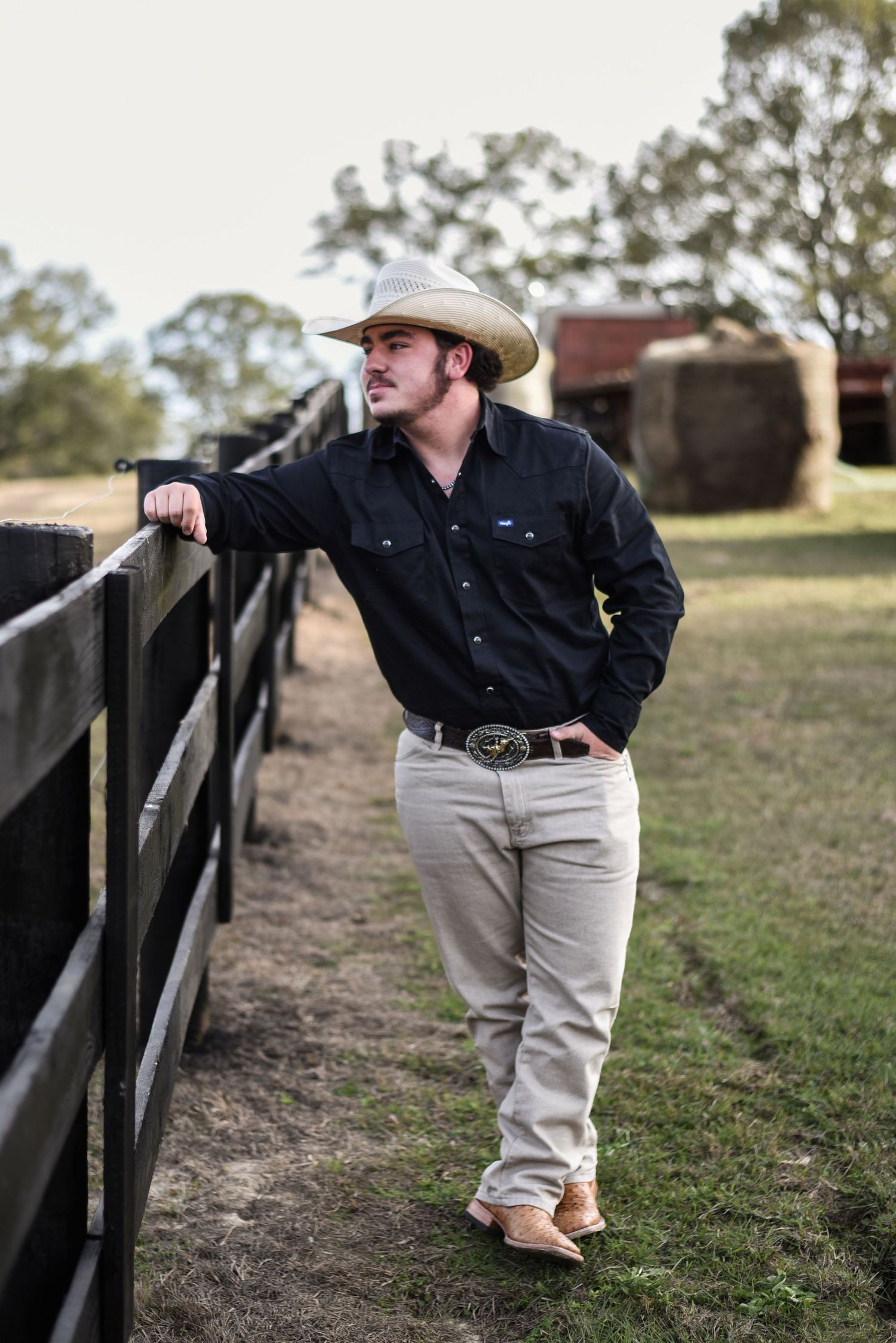 High school senior cowboy leaning against fence in front of barn on a farm in Auburn, Alabama