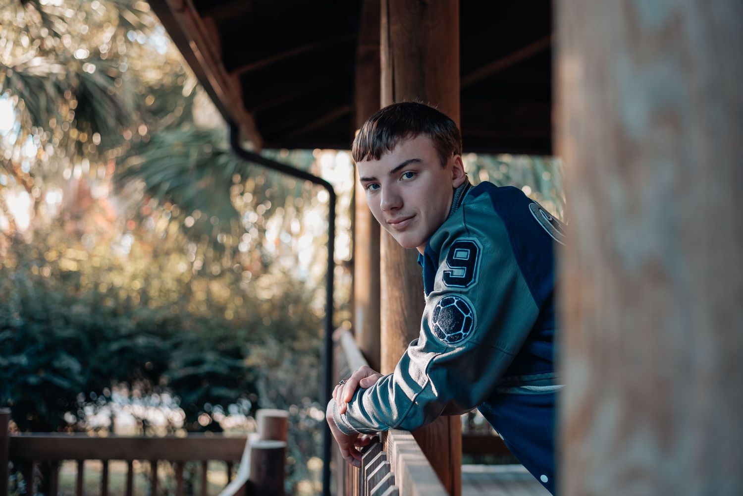 High school senior guy in letterman jacket leaning on porch railing at Blue Heron Nature Center in Ridgeland, South Carolina.
