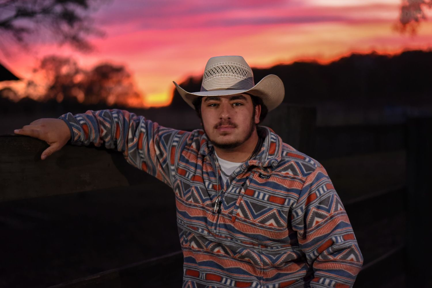 High school senior guy wearing cowboy hat standing at farm fence in front of a colorful sunset in Auburn, Alabama.