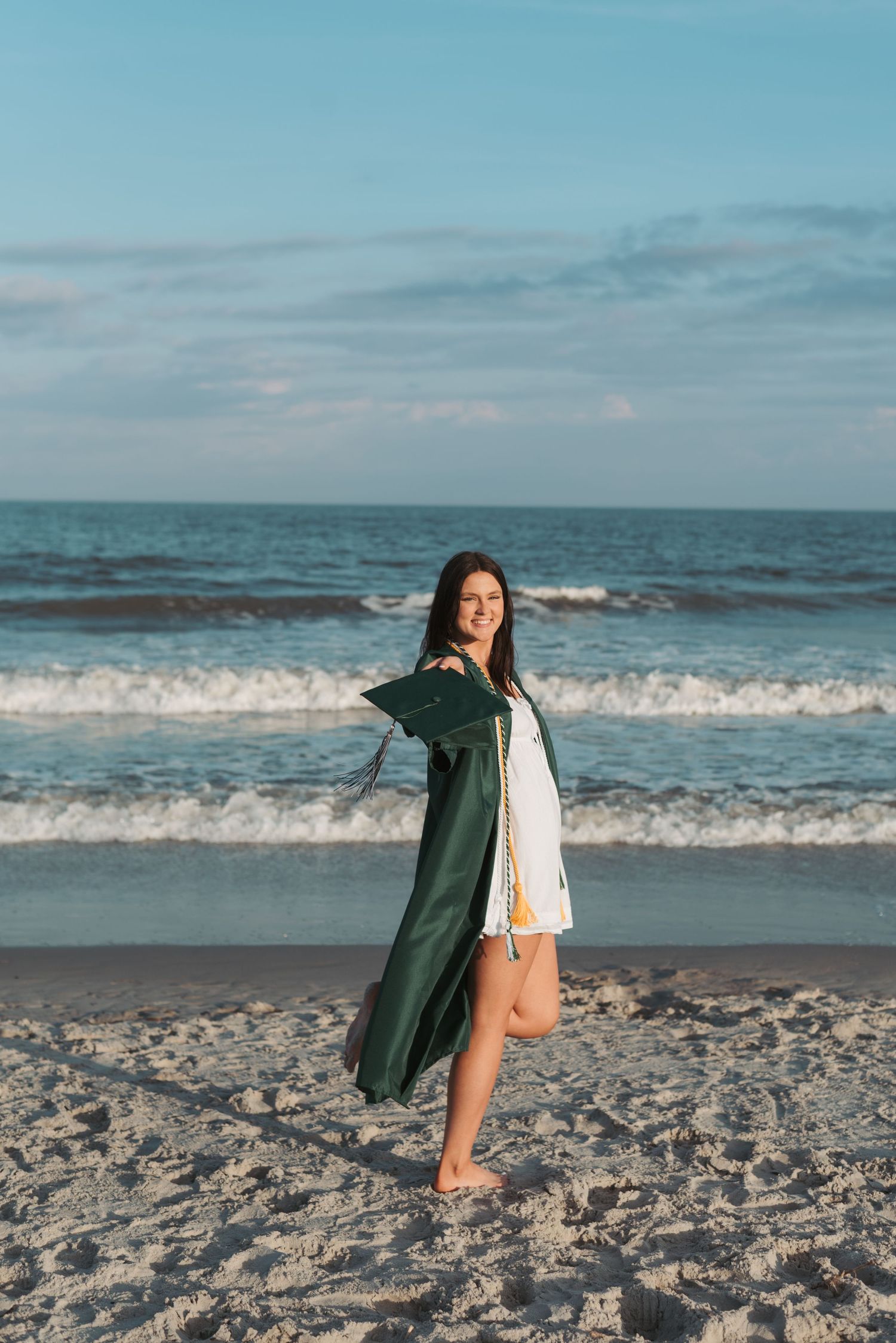 High school senior girl wearing green graduation gown, swinging cap towards camera in front of ocean waves on Hilton Head Island