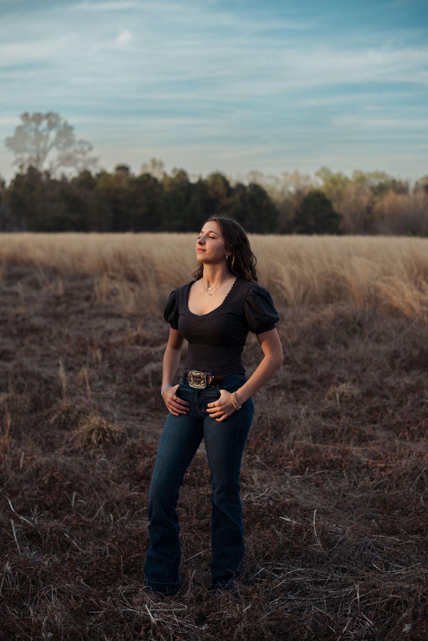 High school senior girl looking up towards setting sun with hands in jean pockets, next to belt buckle, in an open field in Early Branch, SC