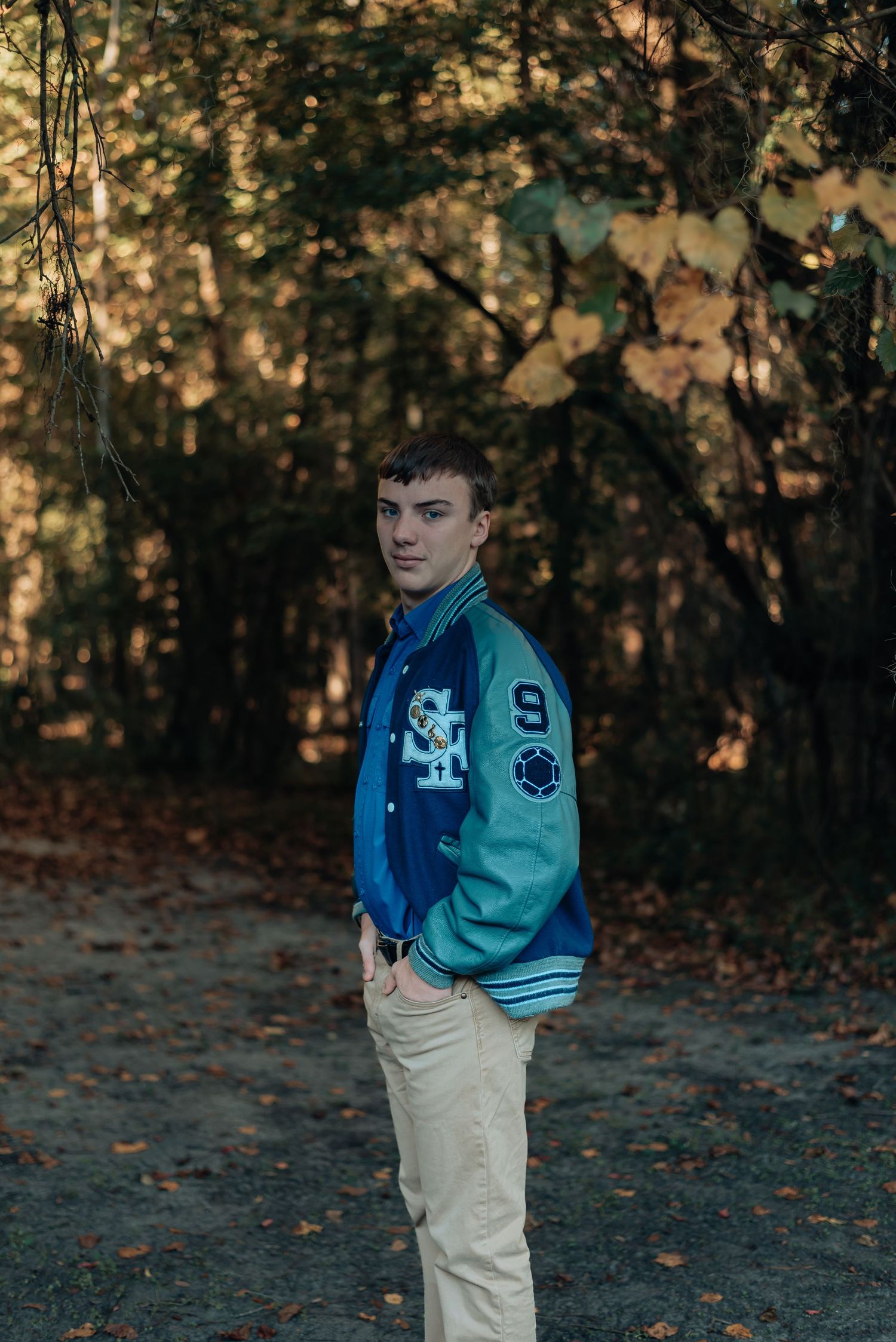 High school senior boy in letterman jacket and khaki pants standing in front of trees at Blue Heron Nature Center Ridgeland, South Carolina