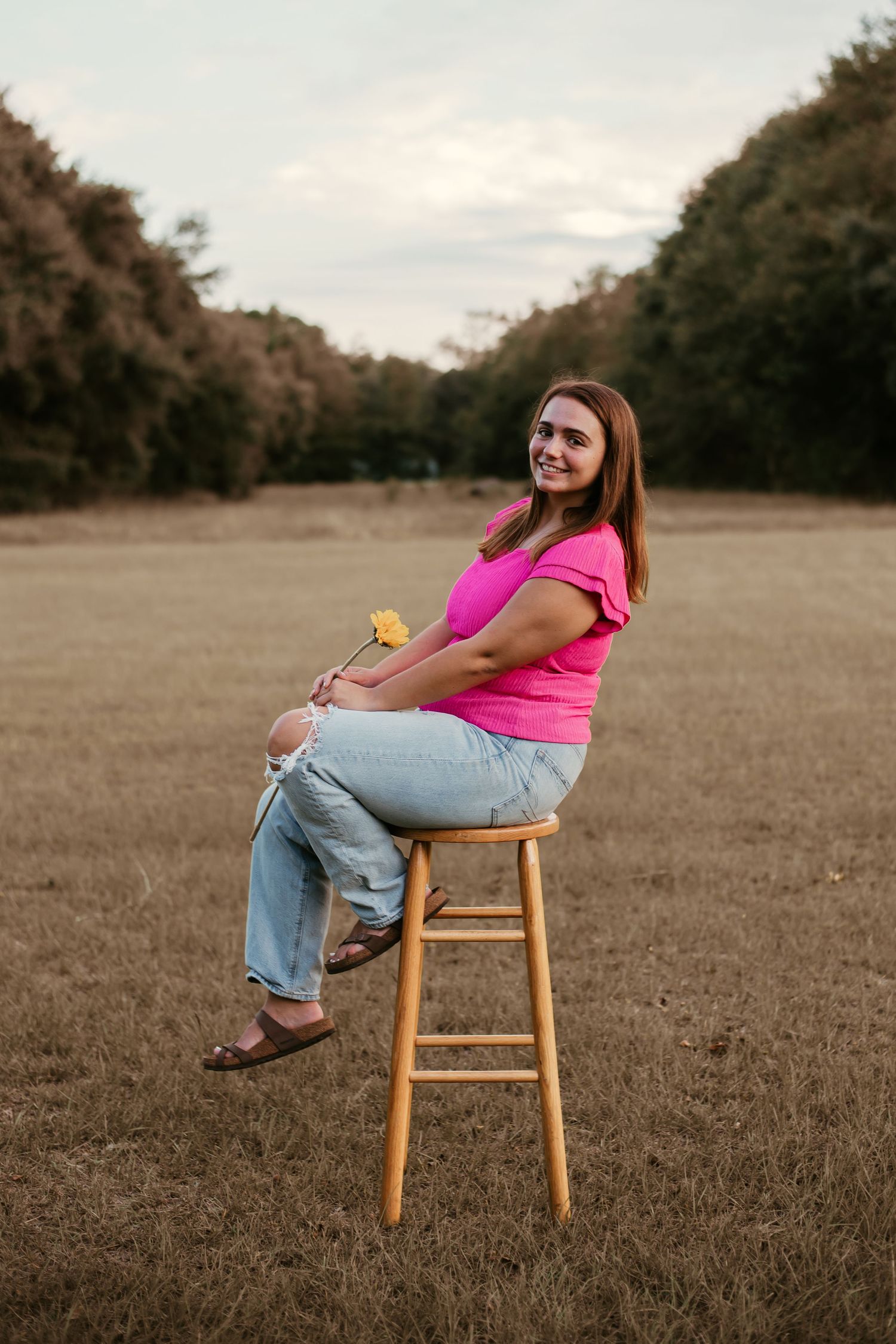 High school senior girl in pink shirt, sitting on stool, holding sunflower in open field in Grays, South Carolina.