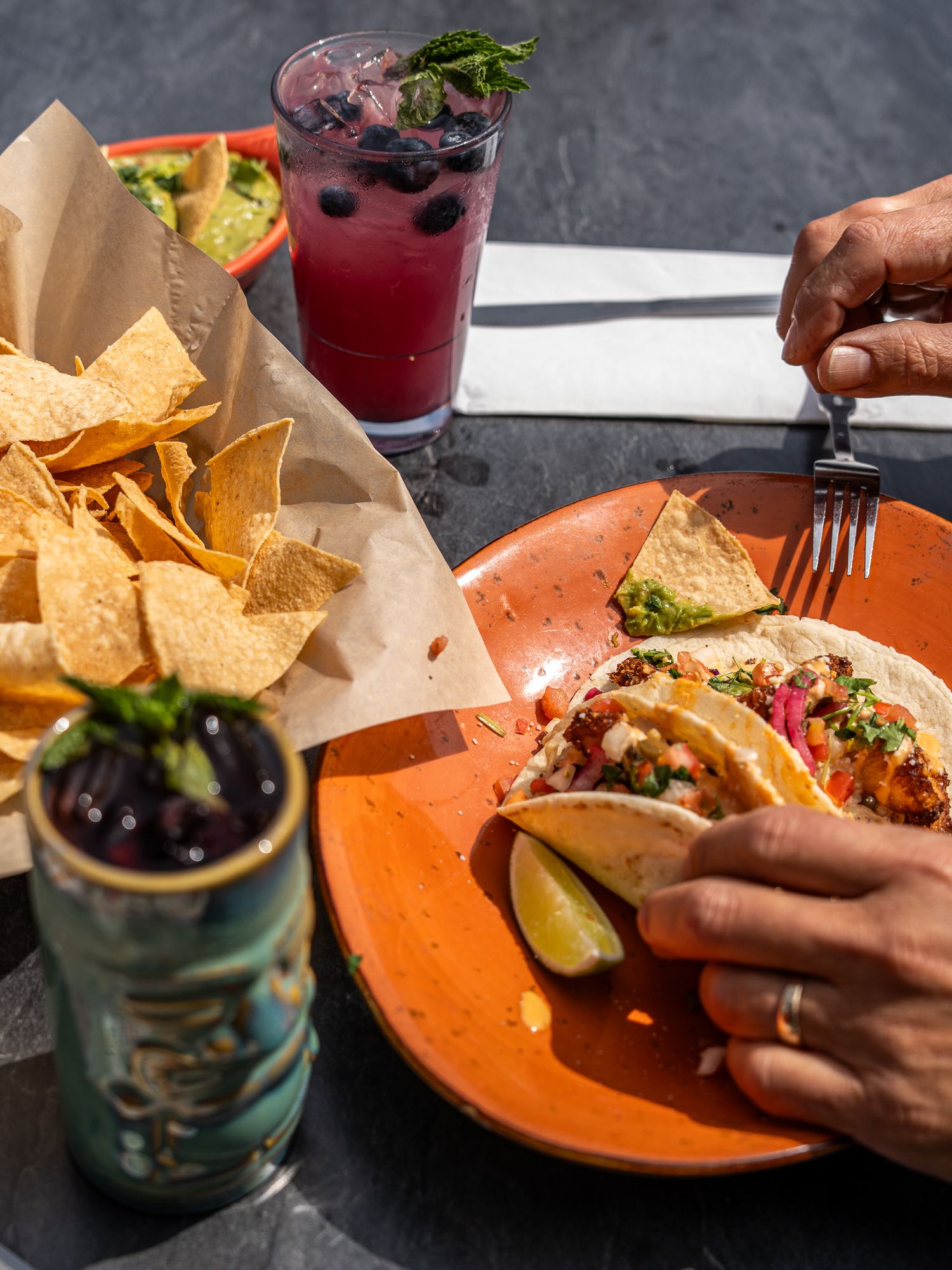 Mexican restaurant table with tacos, tortilla chips, and colorful cocktails garnished with fresh herbs and fruit.