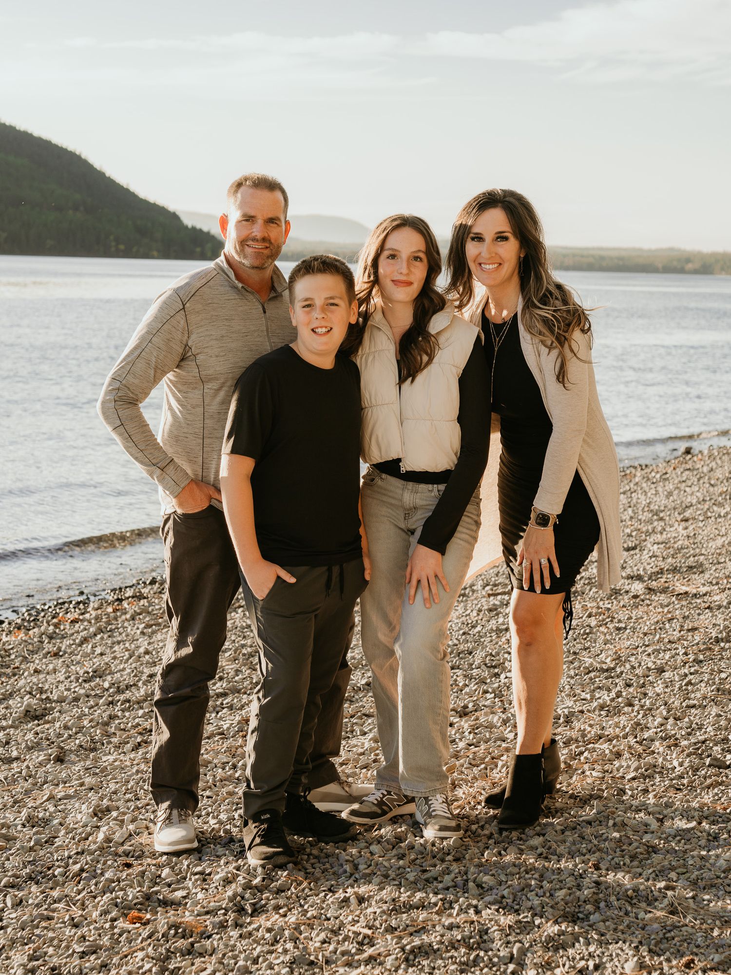 Family of four smiling together on a rocky beach at sunset with mountains and lake in background.