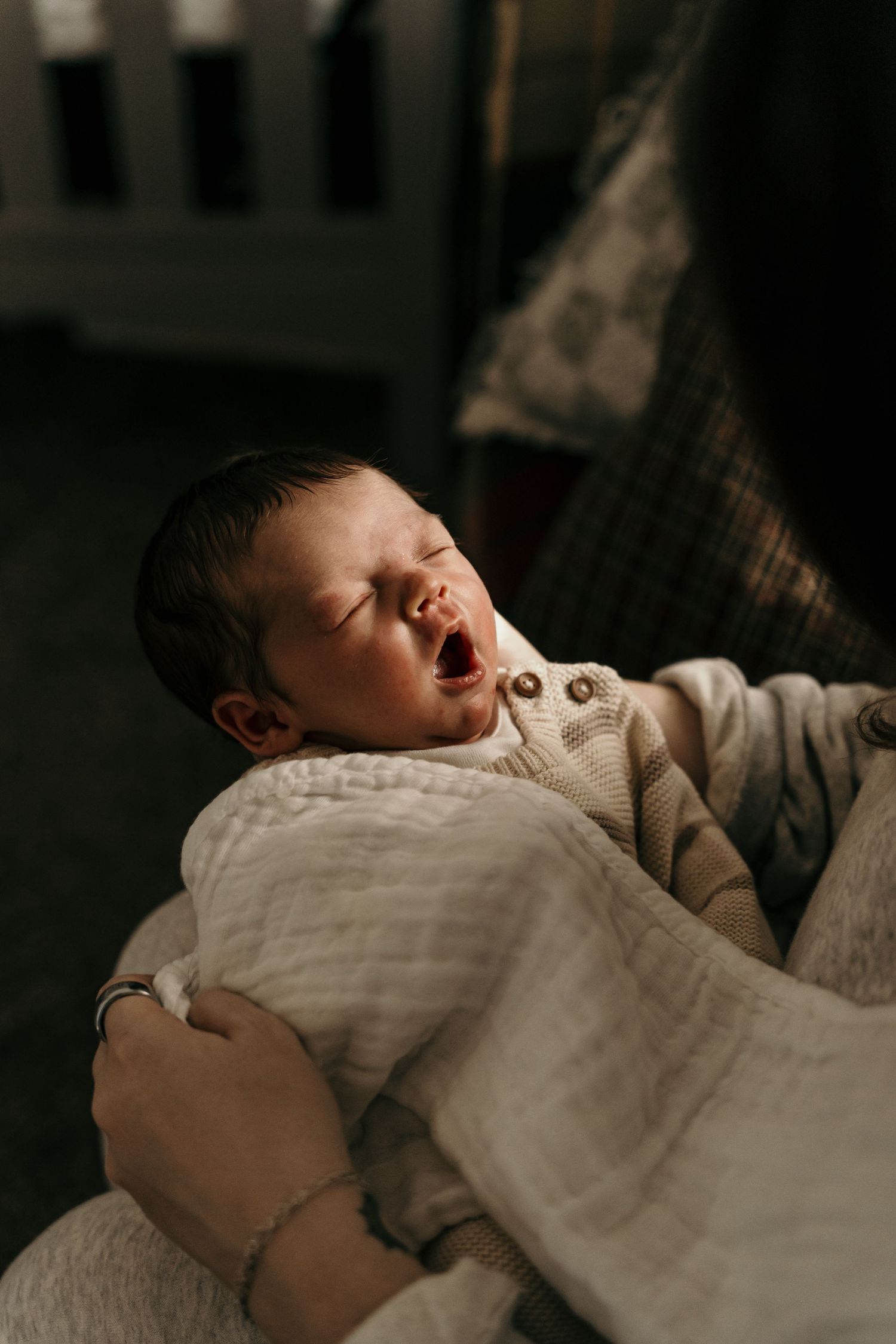 Newborn baby yawning while being held in a white muslin blanket in soft warm light.