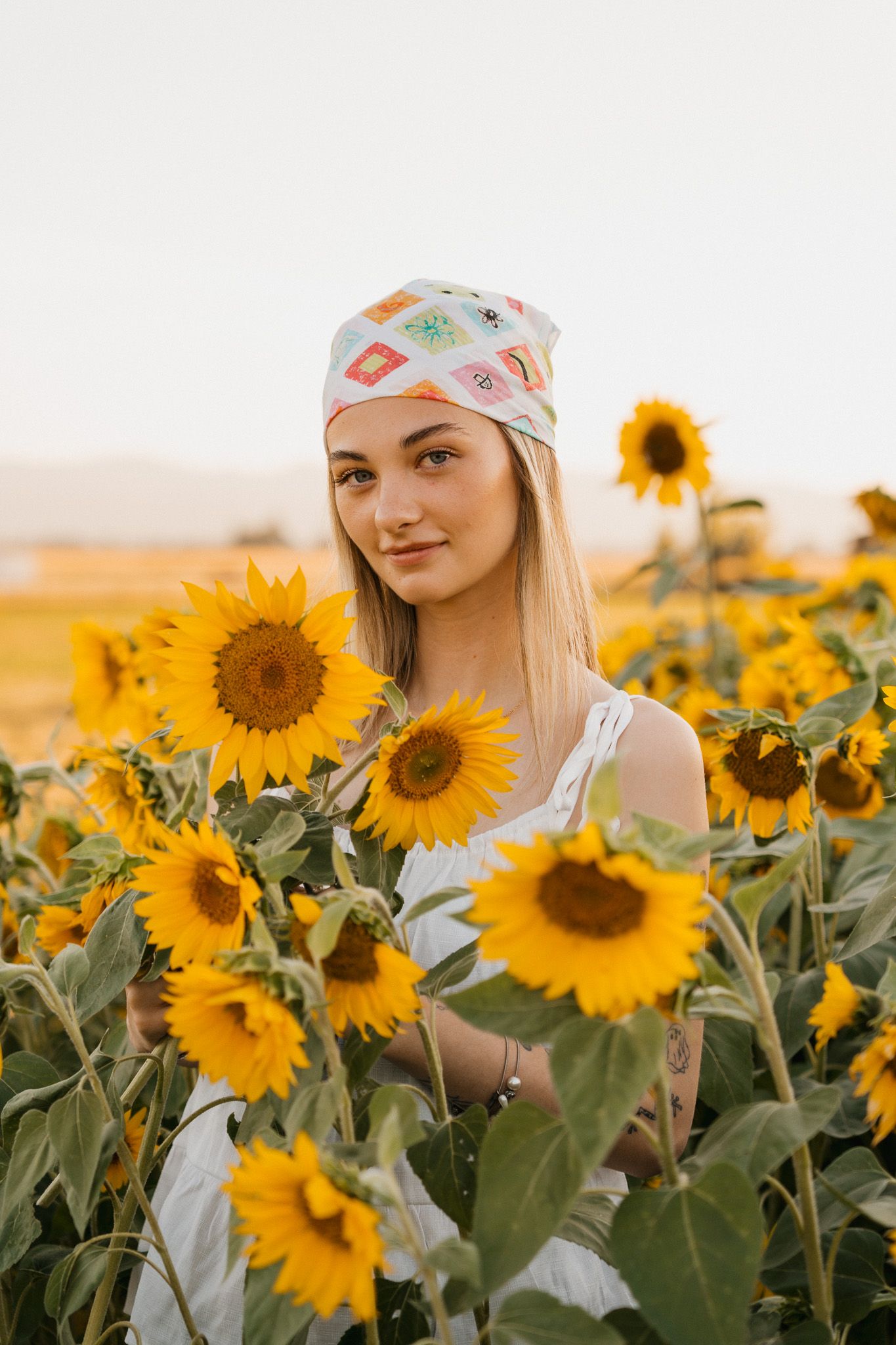 Young woman in white dress and patterned headscarf smiling among vibrant sunflowers in a golden field.