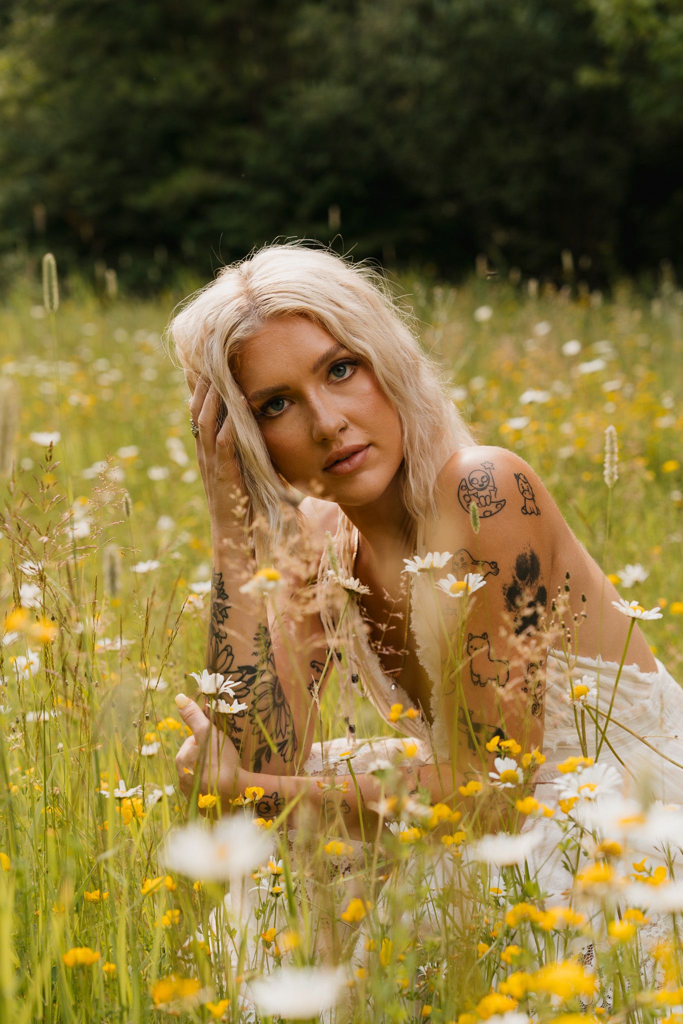 Tattooed blonde woman lying in a wildflower meadow with daisies and yellow flowers, gazing at camera.