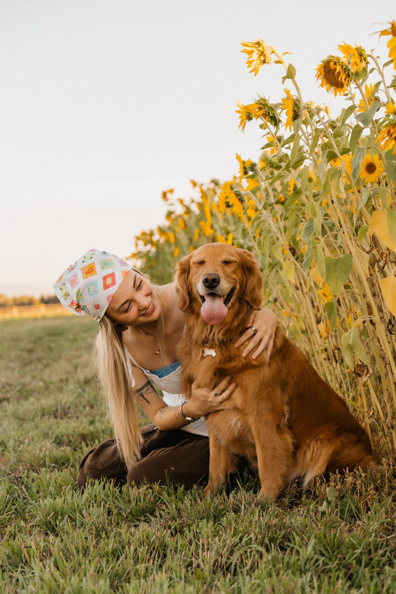Young woman in colorful cap hugging happy Golden Retriever dog beside sunflower field on sunny day.