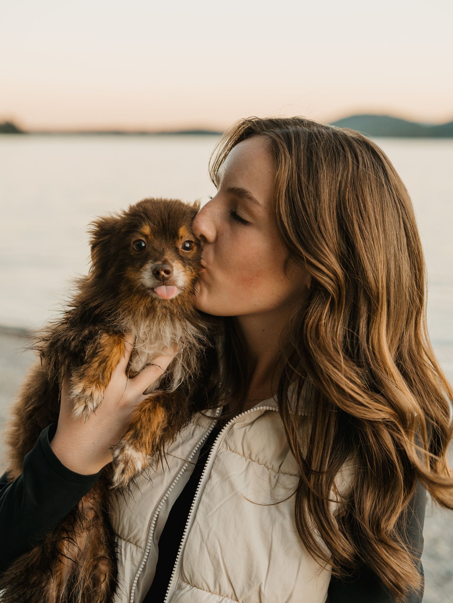 Woman kissing her small brown Pomeranian dog on the cheek at a serene lakeside during golden hour.