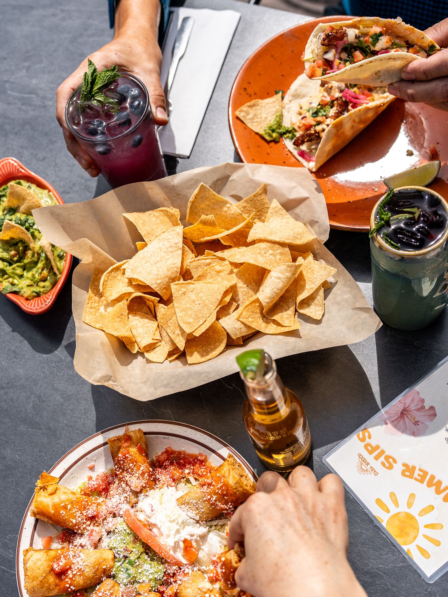 Overhead view of Mexican food spread with tacos, chips, cocktails and beer on gray surface.