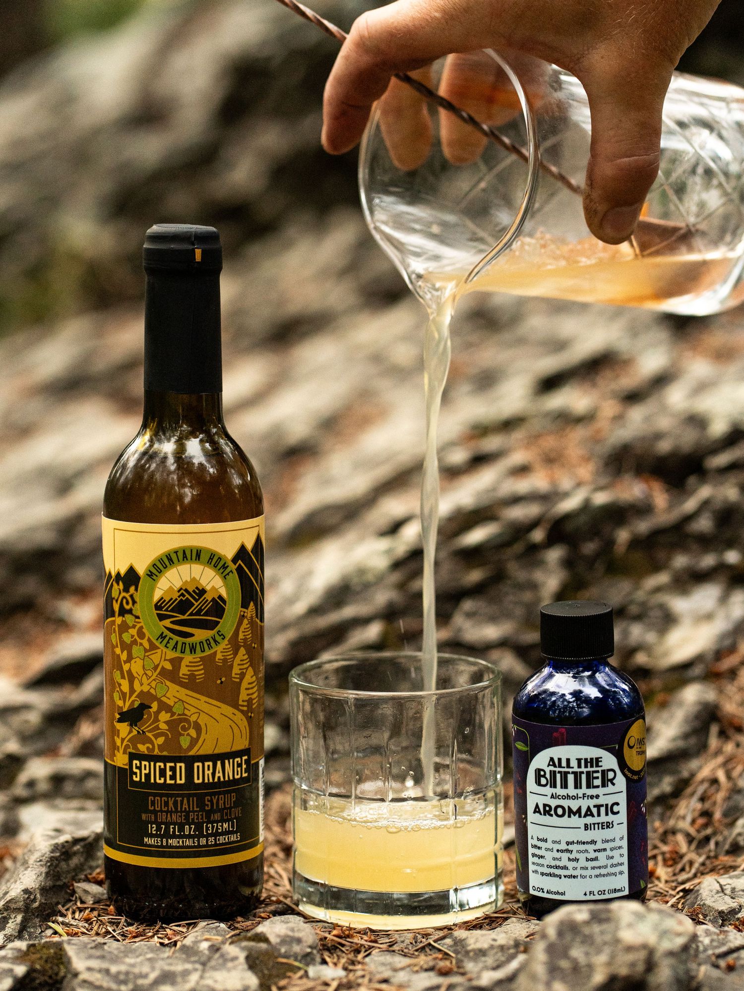 Pouring a cocktail mixture into a glass with orange garnish and small blue bottle on rocky surface.