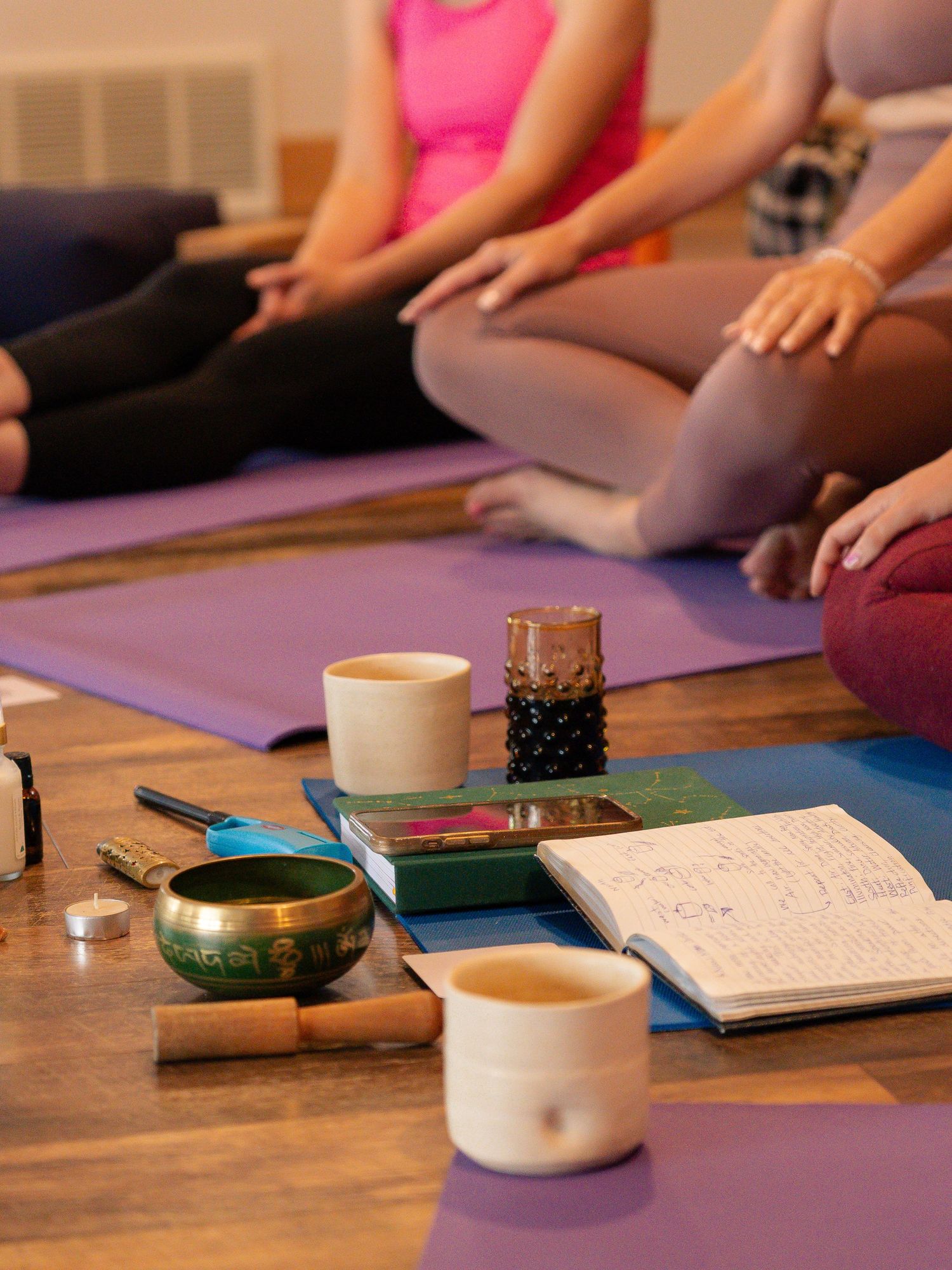 Close-up of meditation setup with singing bowl, tea cups and journals on wooden floor with purple mats.