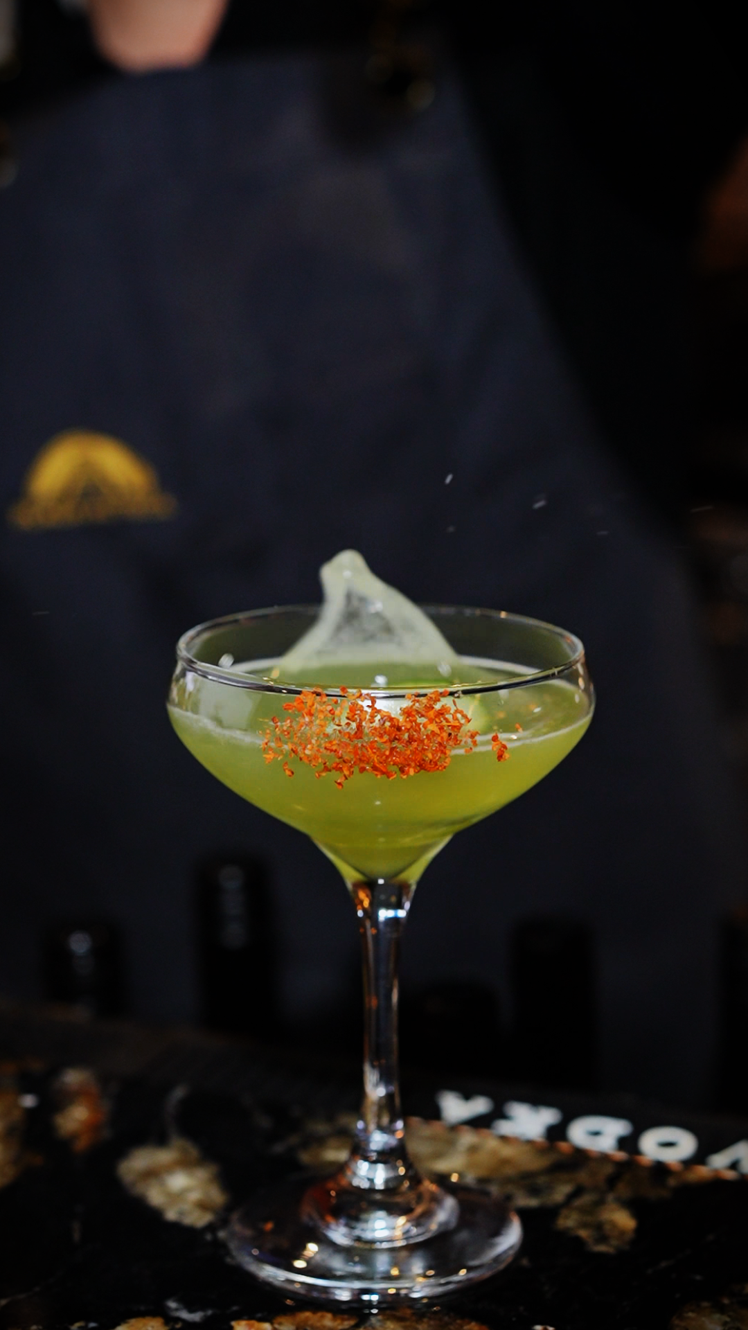 An elegant green cocktail served in a coupe glass garnished with orange tobiko roe and a wasabi foam on top.