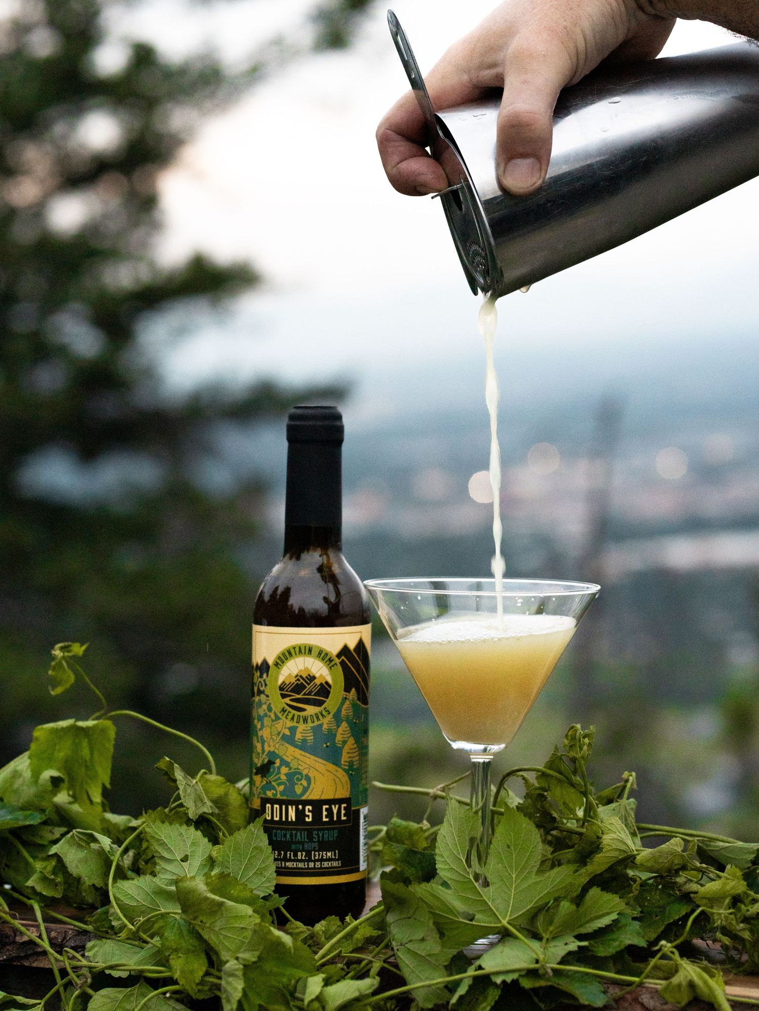 Pouring a yellow cocktail into martini glass with bottle of Lion's Eye liqueur against city backdrop.