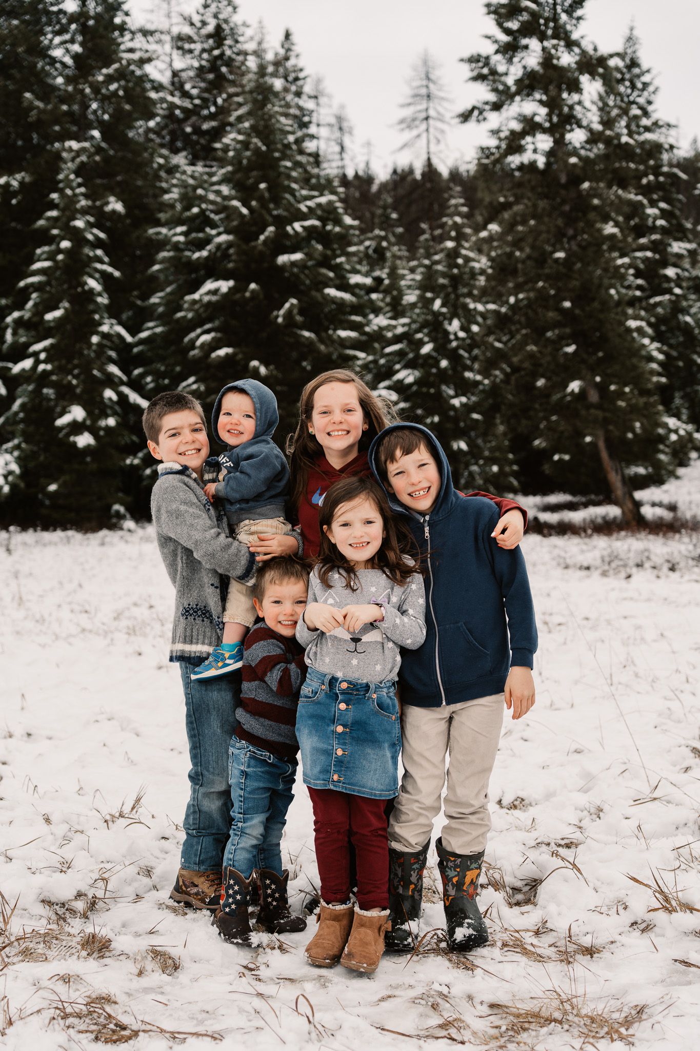 Six children pose together in a snowy forest clearing, wearing winter clothes, smiling for a family photo.