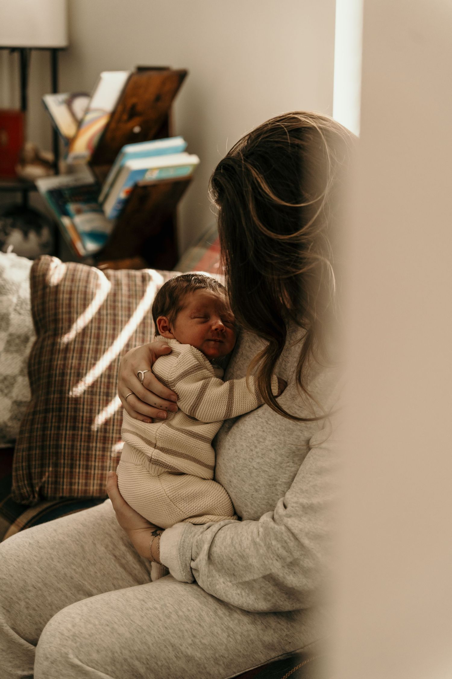 A mother tenderly holds her sleeping newborn baby in soft natural light, sitting near a window in a cozy home.