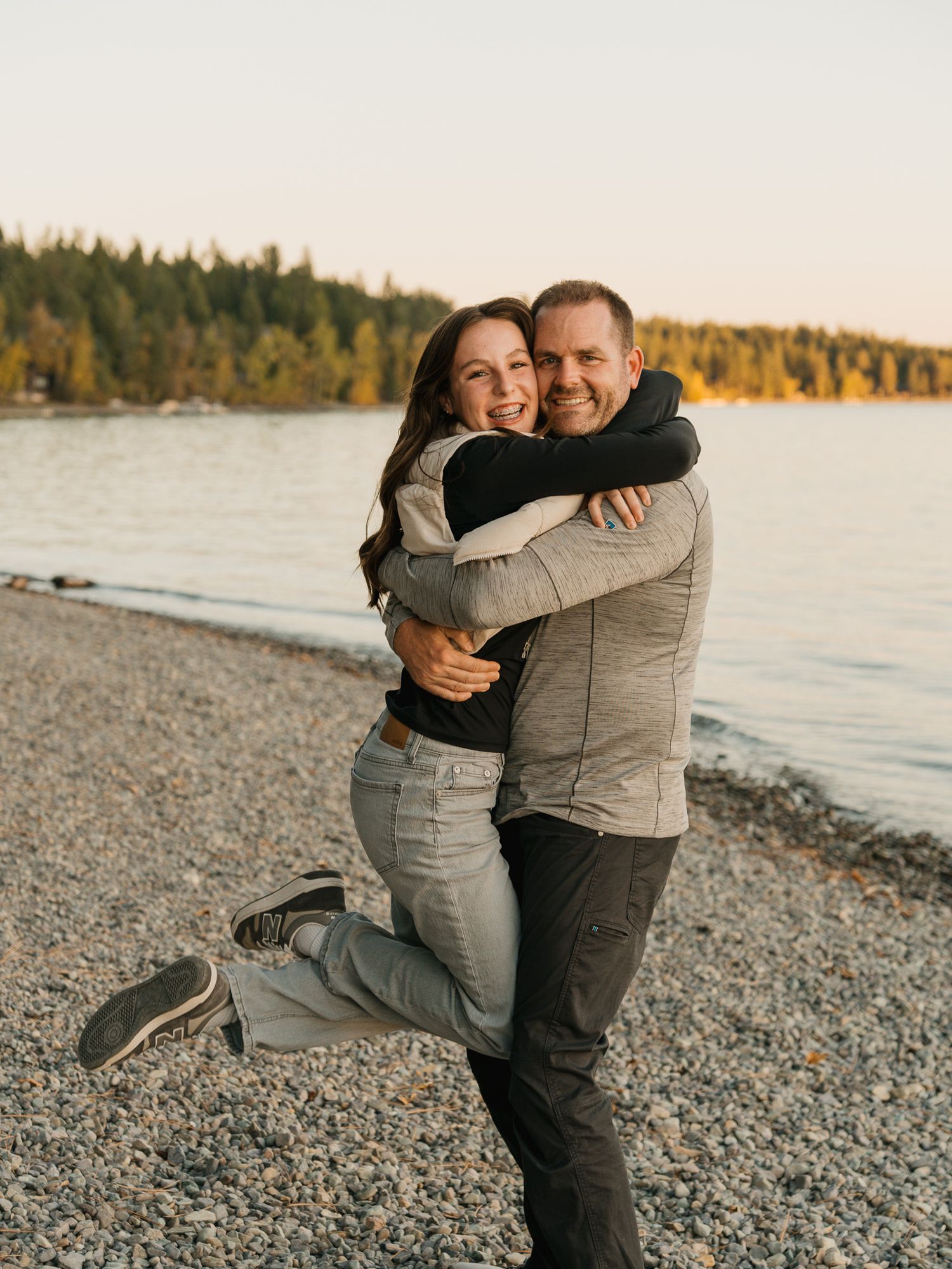 A couple embraces joyfully on a rocky beach at sunset, with a serene lake and forest in the background.