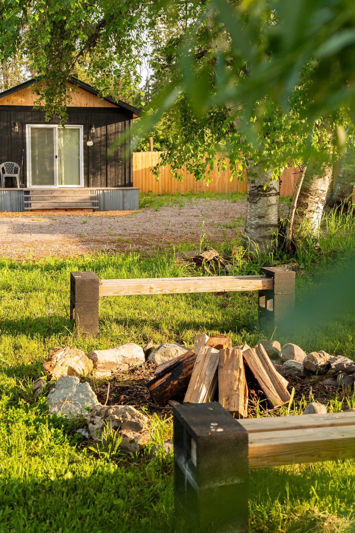 Rustic cabin with fire pit in foreground surrounded by birch trees and green foliage.