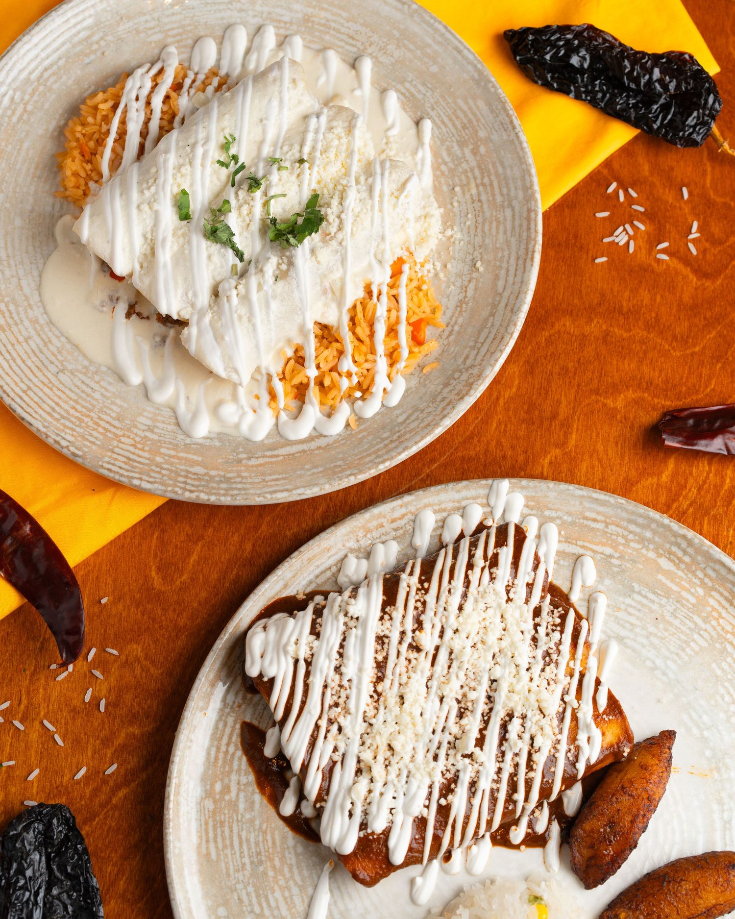 Two Mexican-style plates drizzled with white sauce and garnished with dried chiles on an orange surface.
