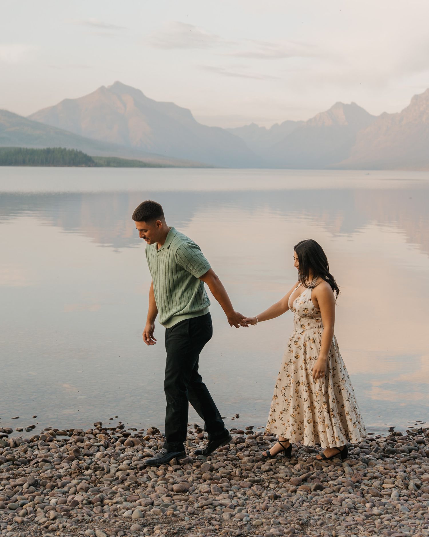 Couple holding hands walking along rocky lakeshore with mountain reflections at sunset.
