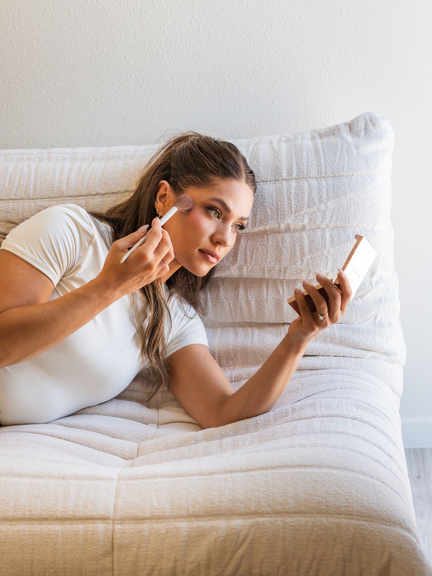 Person applying makeup while looking in handheld mirror while relaxing on white couch.