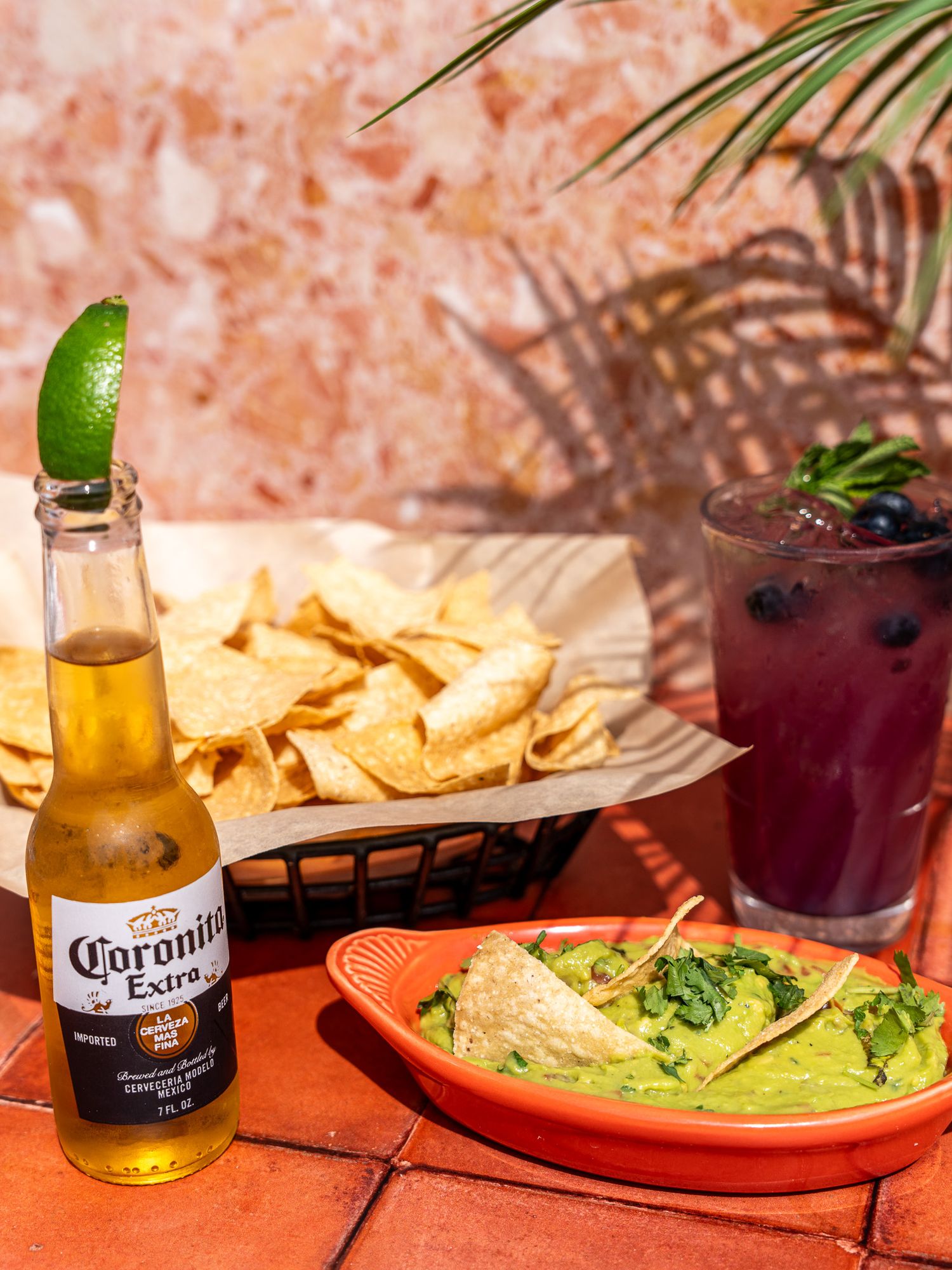 Corona beer bottle with lime, berry cocktail and guacamole served with tortilla chips on coral table.