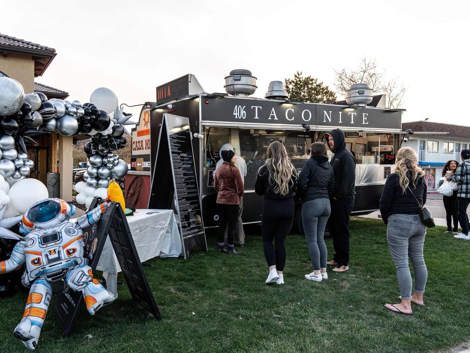 A taco food truck with black balloons and customers waiting in line on grass.