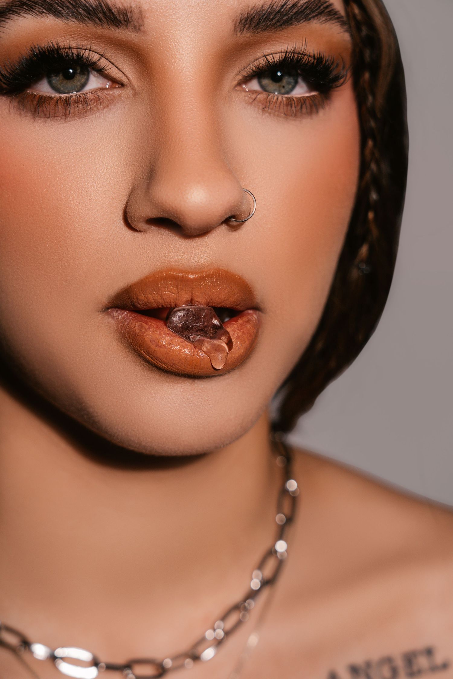Close-up beauty shot showcasing glossy brown lipstick and dramatic eye makeup with silver chain necklace.