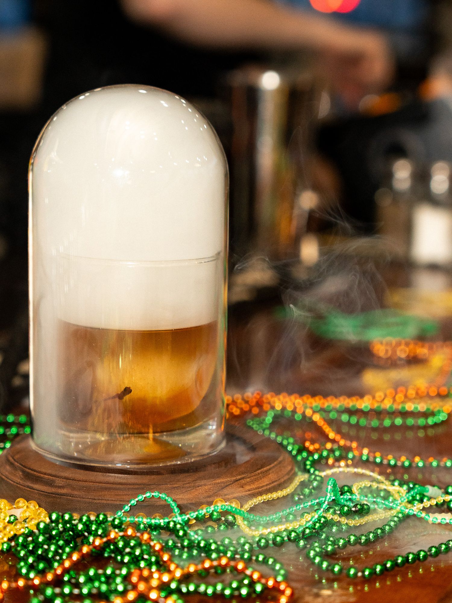A smoky cocktail under a glass dome surrounded by festive green and gold Mardi Gras beads.