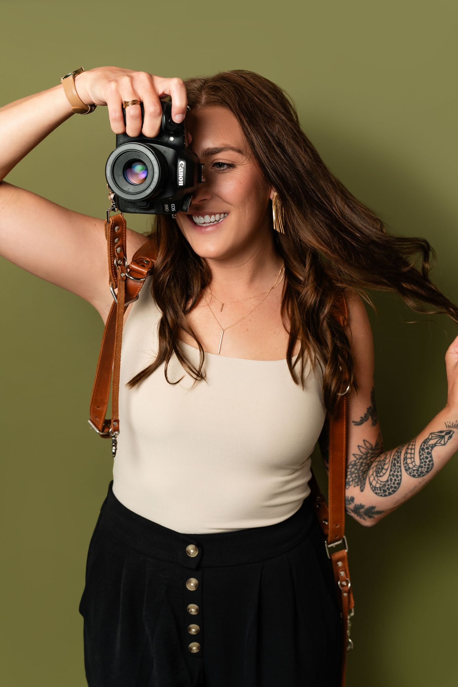 Photographer with long brown hair wearing white tank top and black skirt smiles while taking picture against olive green background.