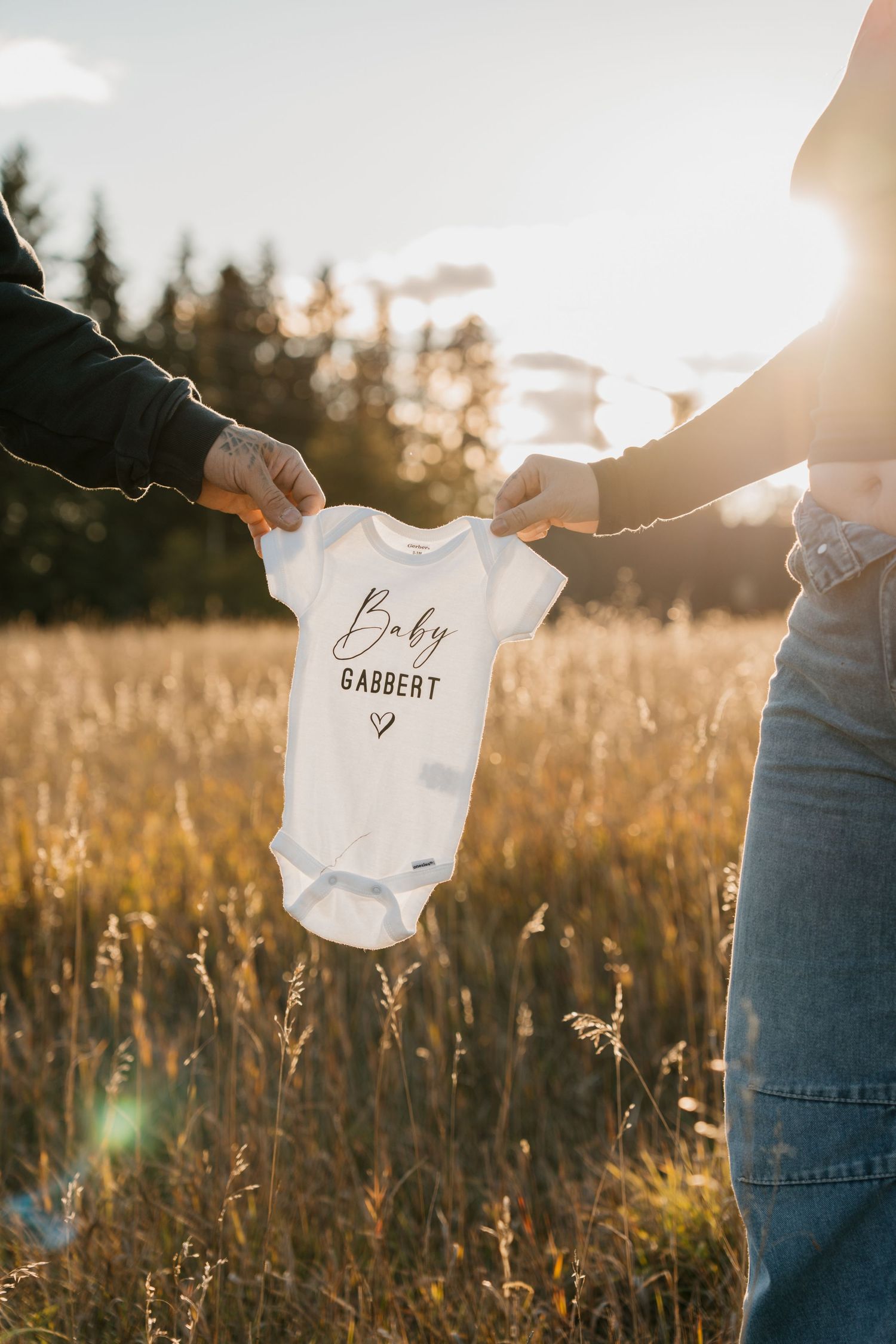 Couple holding a white Baby Gabbert onesie in a golden sunlit field, announcing their pregnancy.
