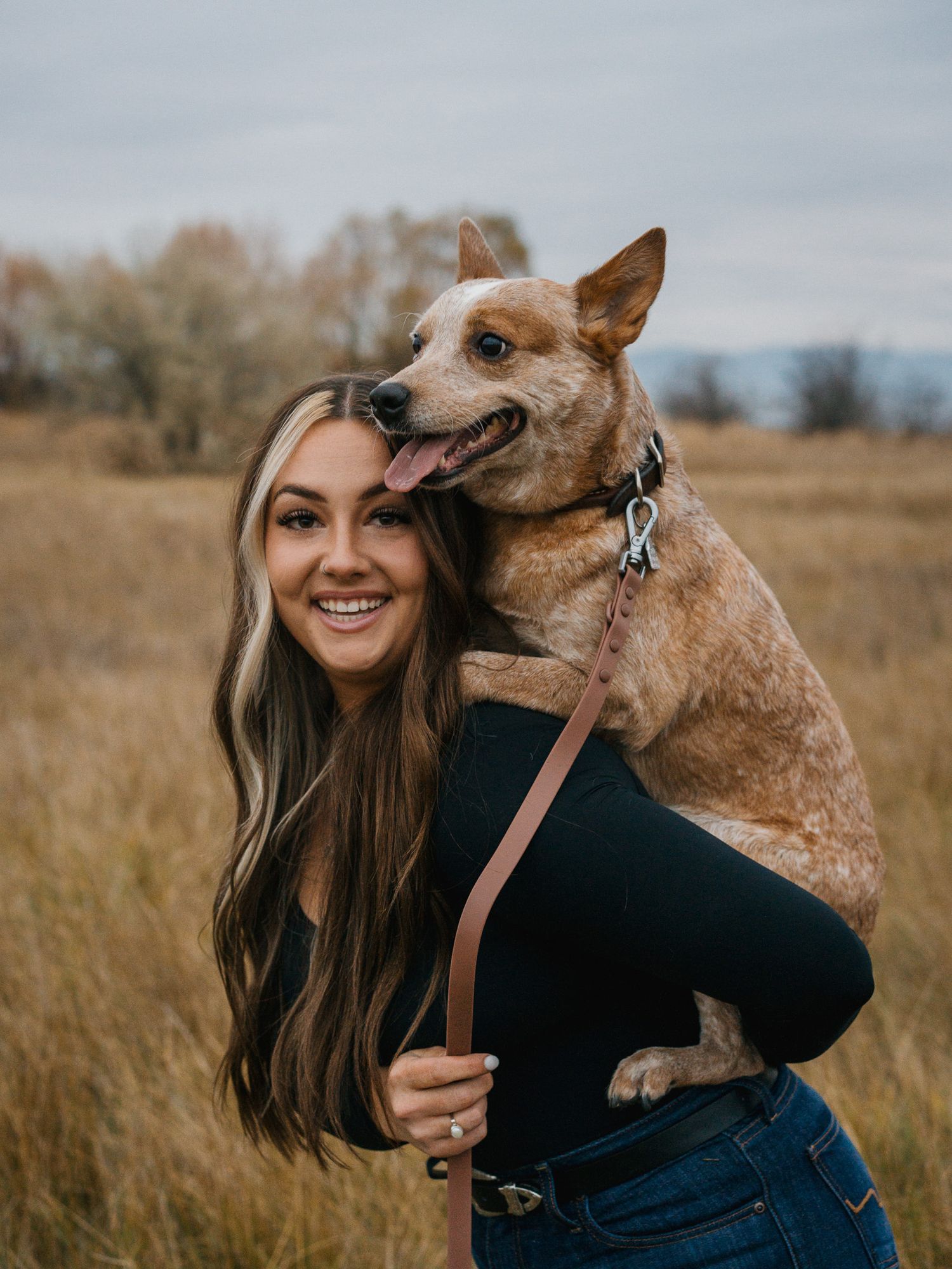 A smiling woman with long brown hair holds an Australian Cattle Dog on her shoulders in an autumn field.